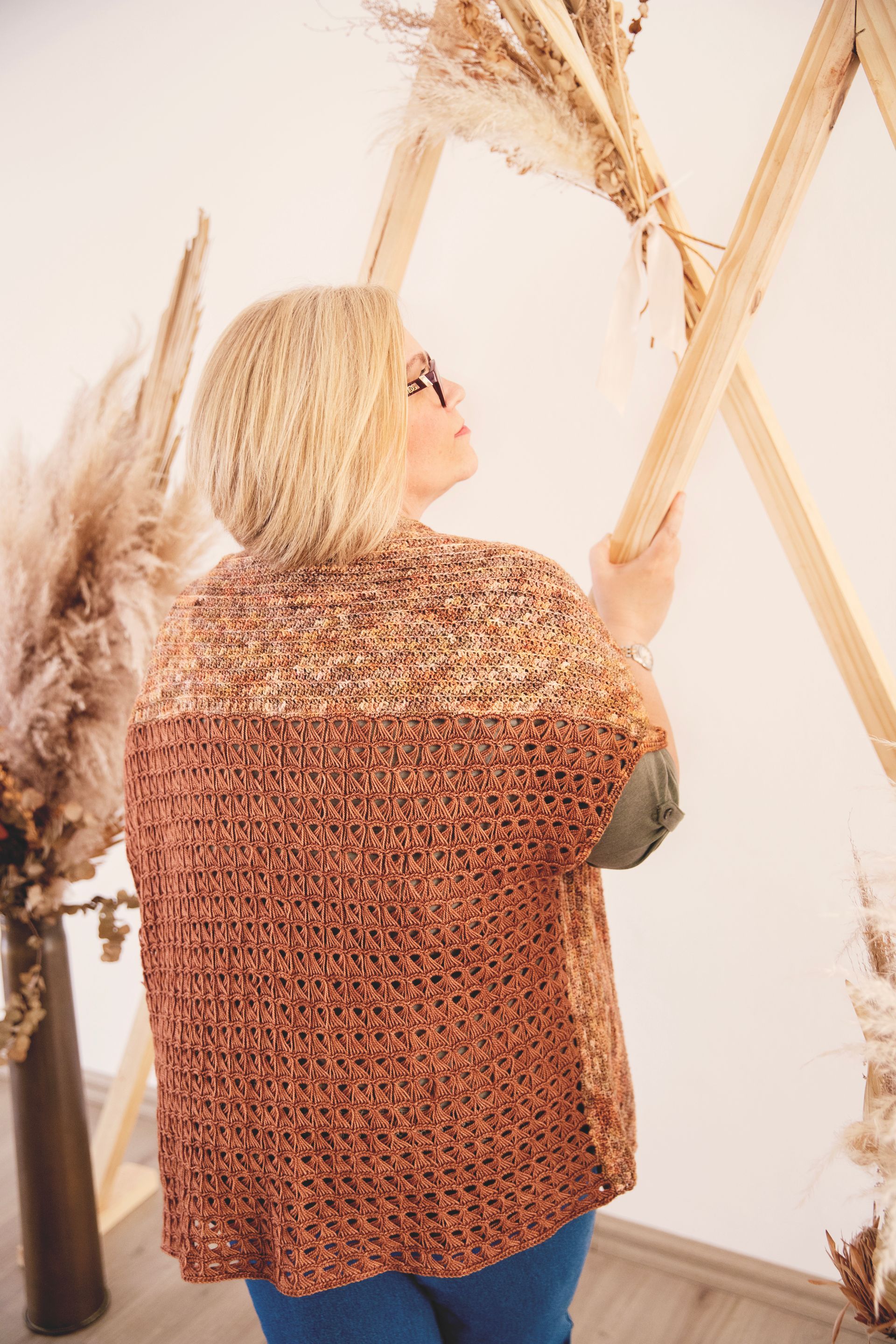 Woman with blonde hair, wearing a brown sweater, arranging pampas grass on a wooden frame.