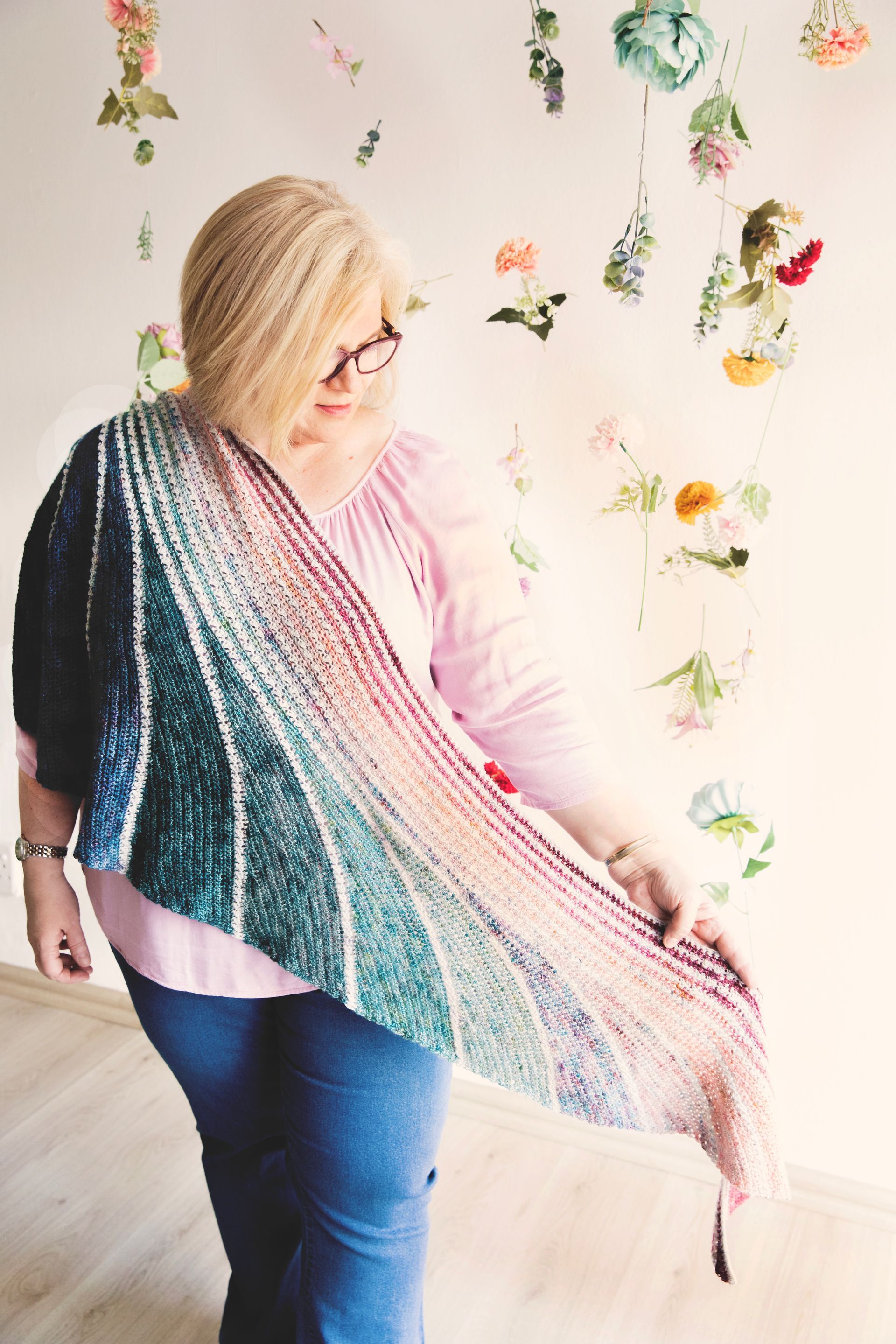 Woman in glasses and pink shirt holding a colorful crocheted shawl indoors, flowers in the background.