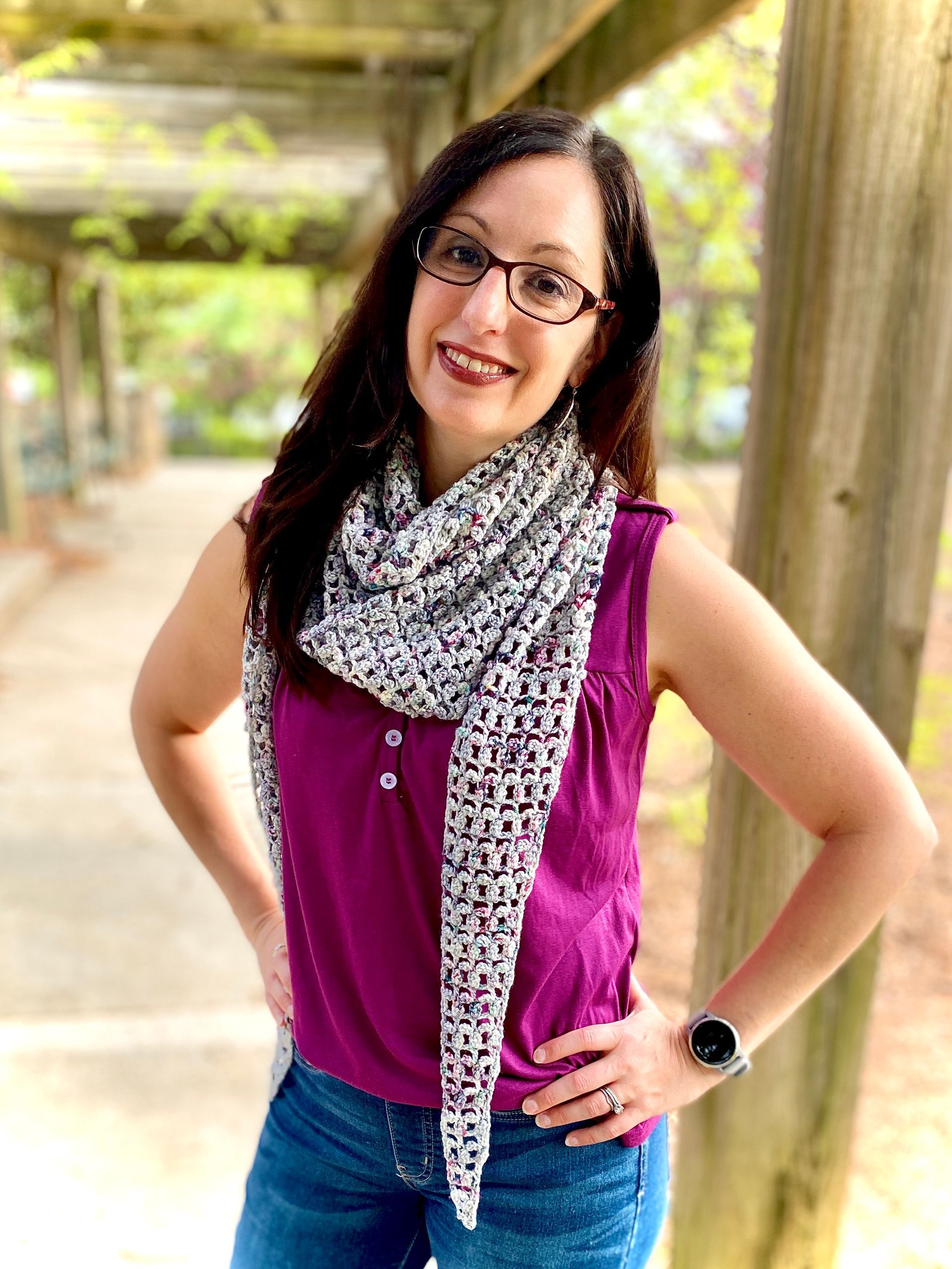 Woman in glasses and scarf poses outside, wearing a purple top and jeans.
