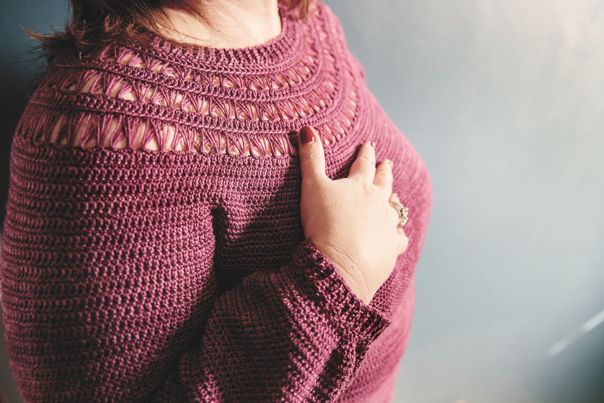 Woman in maroon sweater, hand on chest. Sweater has textured and lacy details.