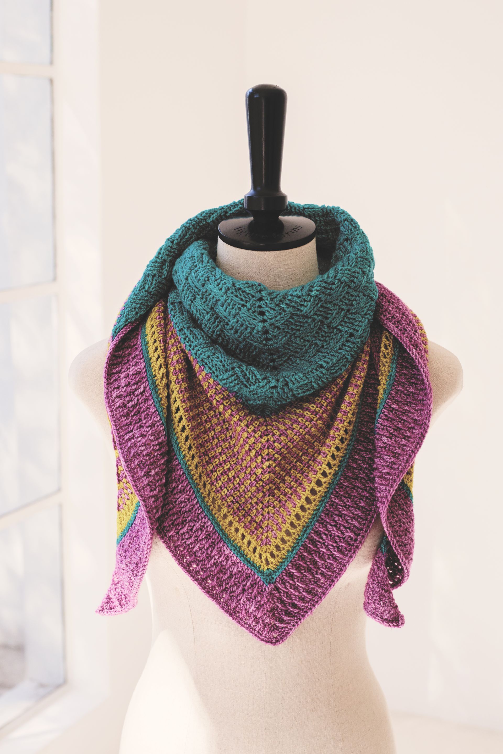 A crocheted triangular shawl in shades of teal, purple, gold, and brown, displayed on a mannequin.