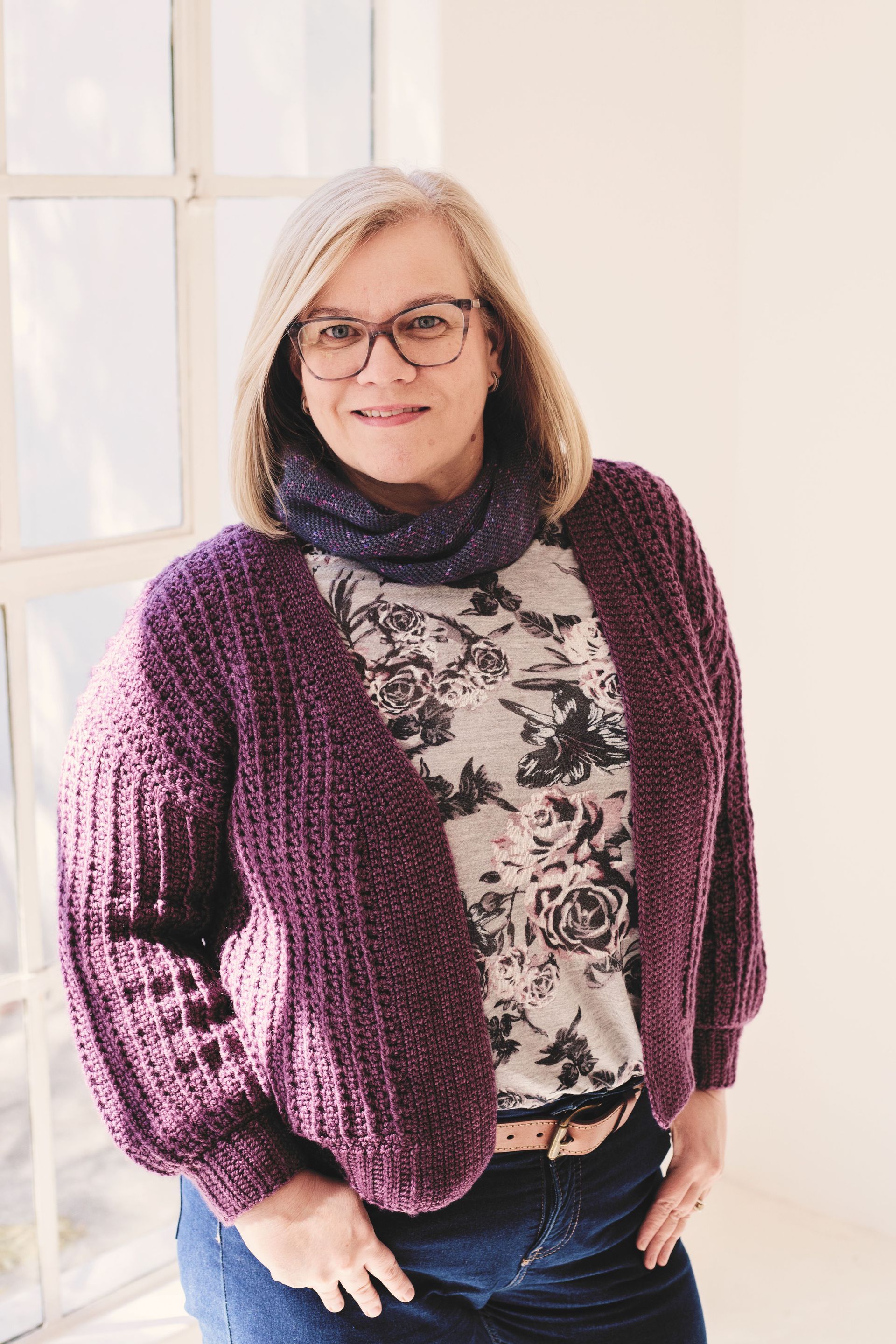 Woman in glasses wearing a purple cardigan, floral shirt, and dark scarf, smiling by a window.