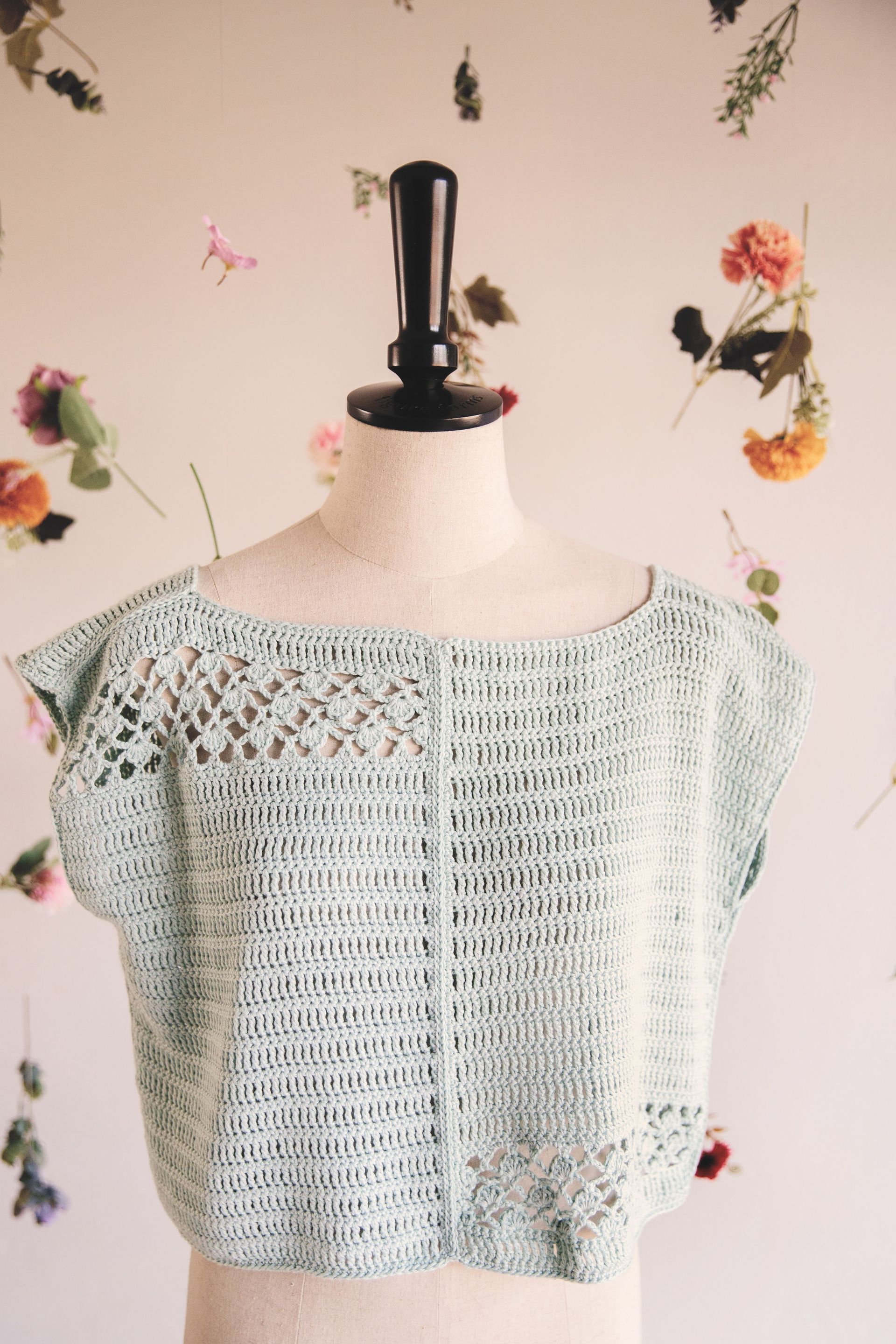 Cropped light blue crocheted top on a mannequin against a backdrop of flowers.
