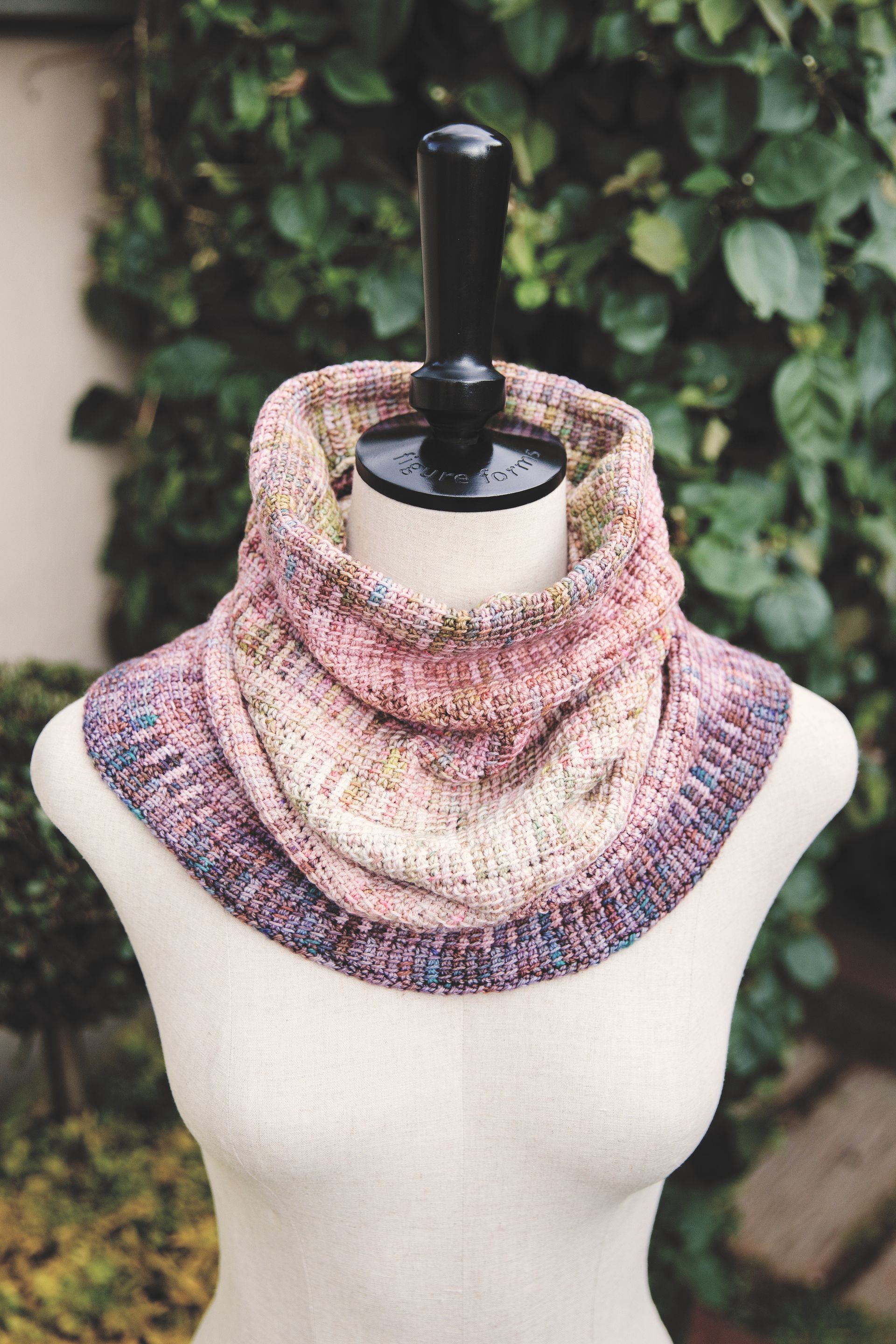 Knitted cowl in pinks and purples, on a mannequin in front of green foliage.