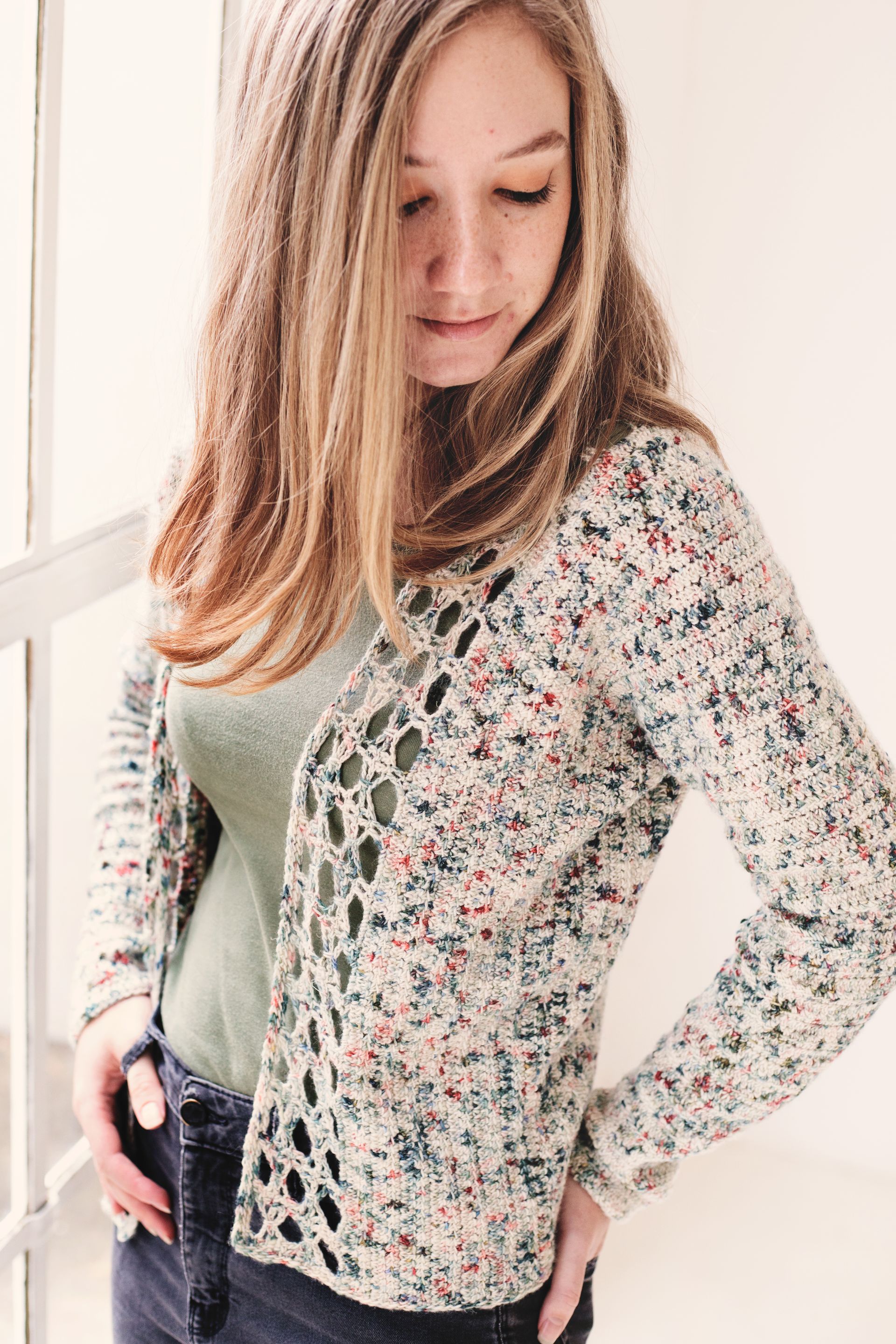 Woman in speckled cardigan, standing indoors near a window, hands in pockets, looking down.