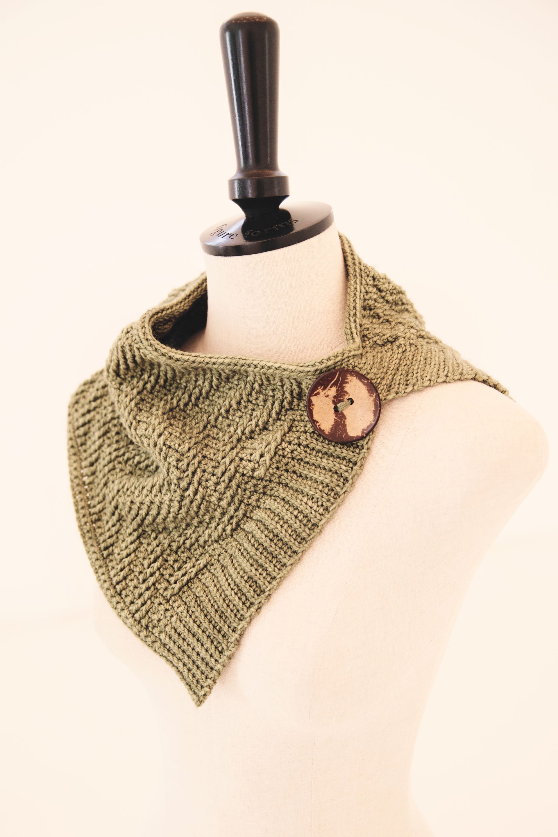 Green knitted cowl with a wooden button, displayed on a mannequin.