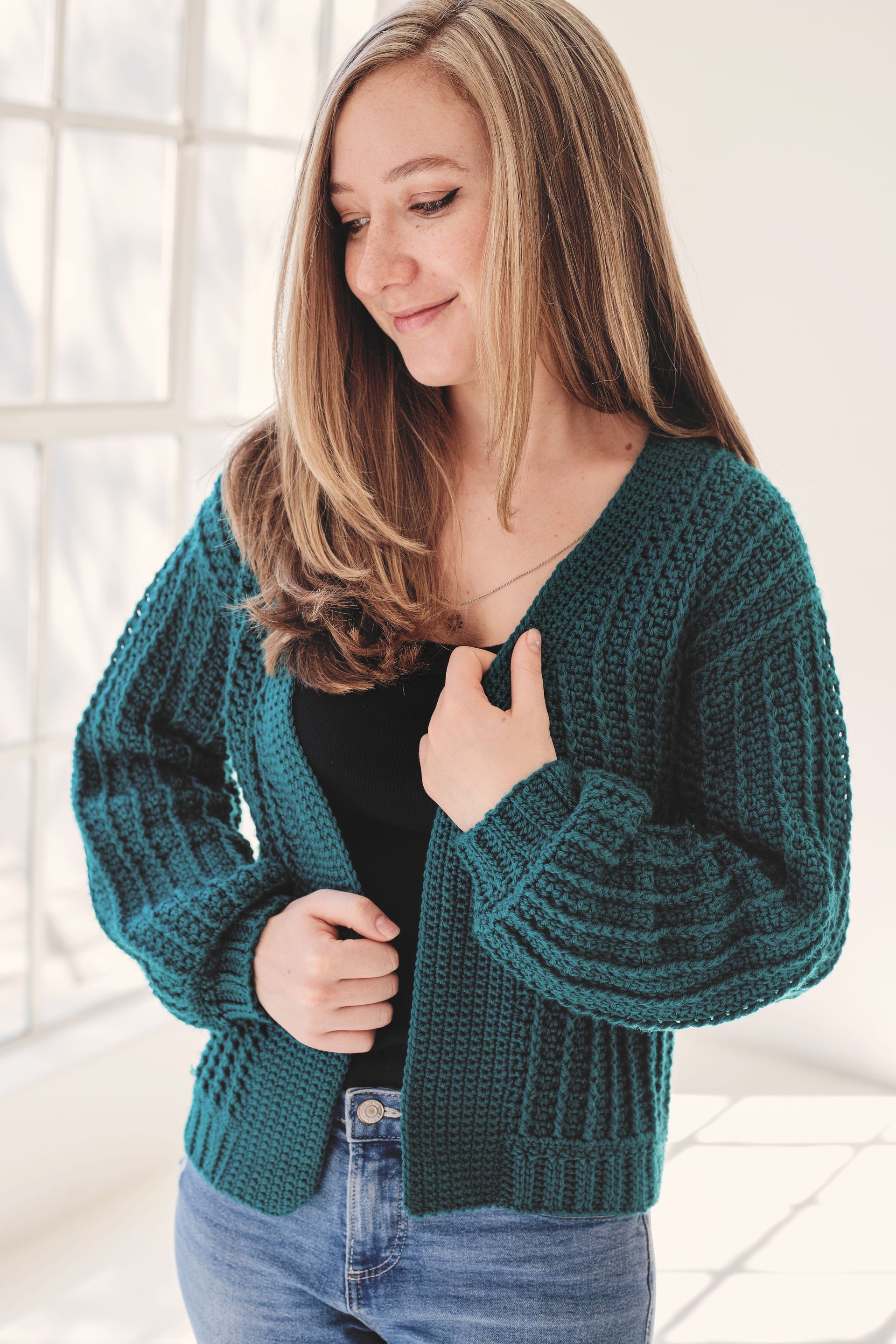 Woman wearing a teal crocheted cardigan, smiling, in front of a window.