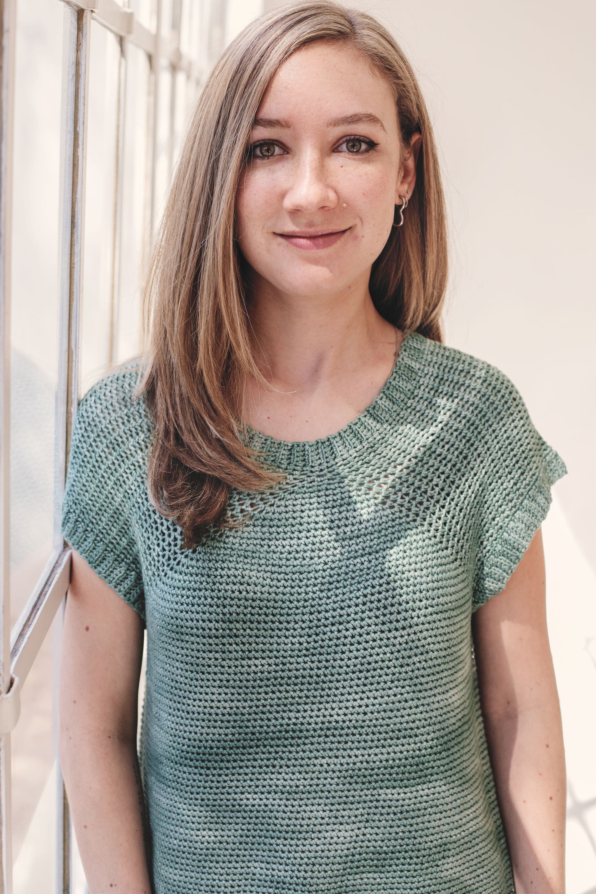 Woman with long blonde hair wearing a green top, standing by a window.