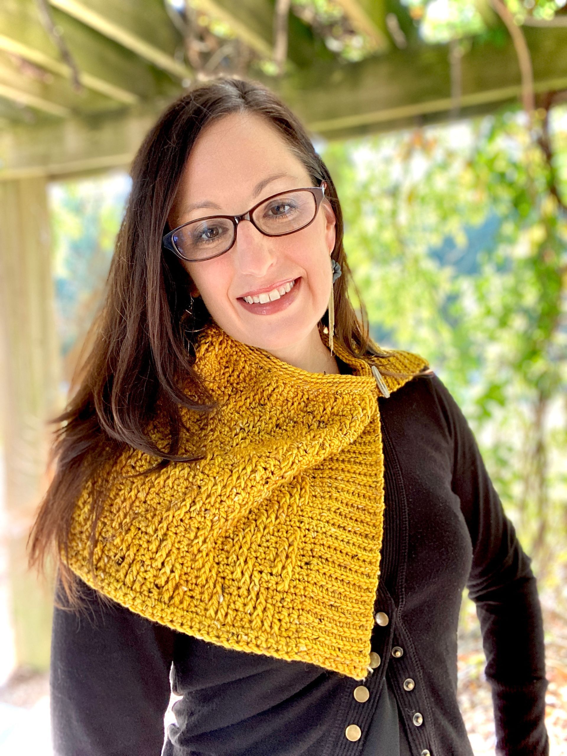 Woman with glasses smiles, wearing a mustard-yellow cowl, black sweater, outdoors.
