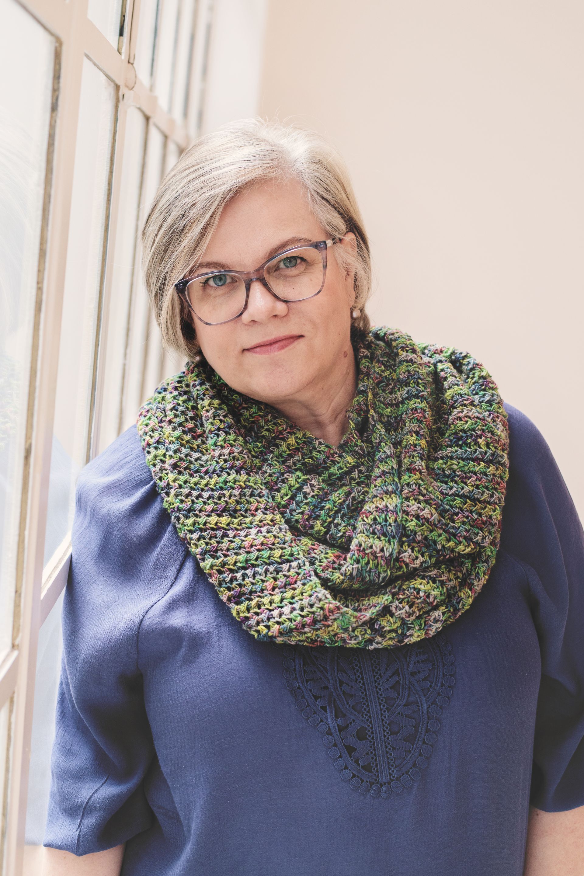 Woman with glasses and gray hair wearing a blue sweater and green and brown cowl leans against a window.