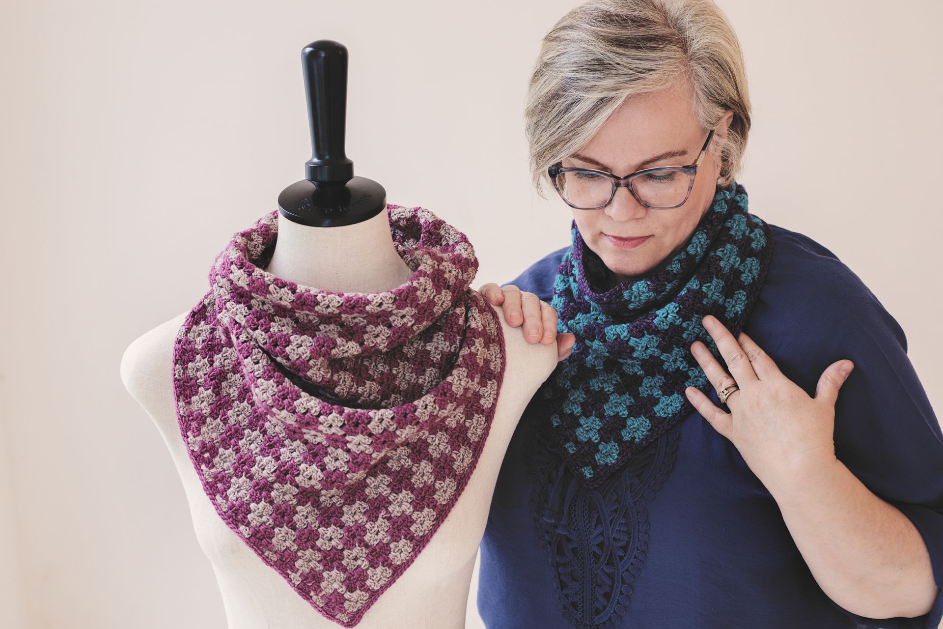 Woman with glasses examines a blue and black patterned cowl; a maroon cowl is displayed on a mannequin.