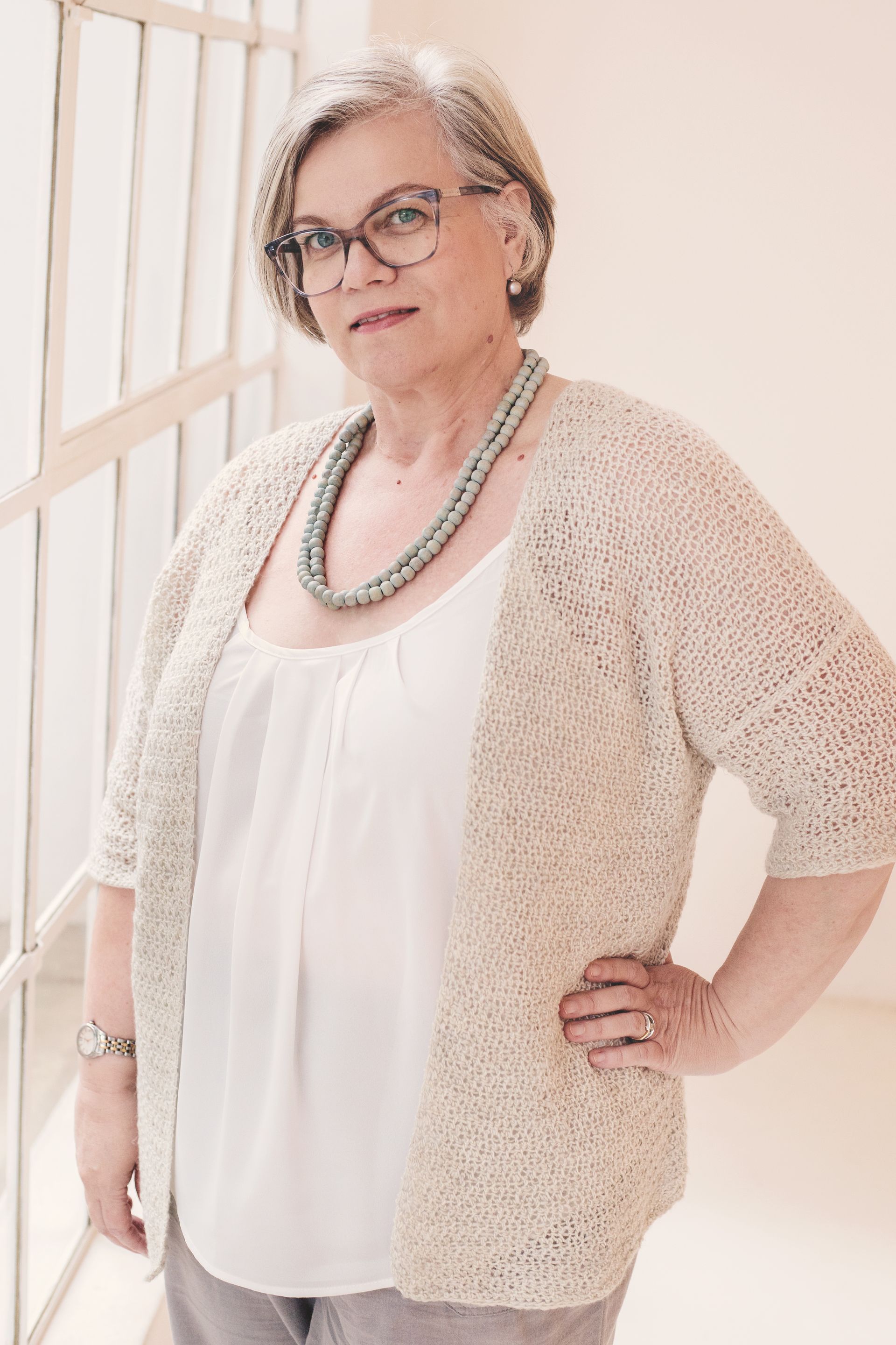 Woman in glasses, standing, hand on hip, by a window; wearing a sweater, white shirt, and necklace.