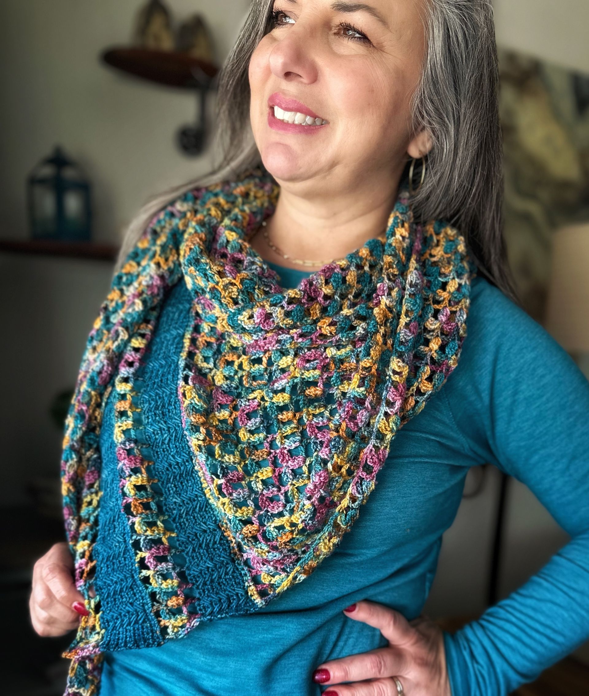 Woman wearing a colorful crocheted shawl smiles, standing indoors. She wears a blue shirt, arms at her side.