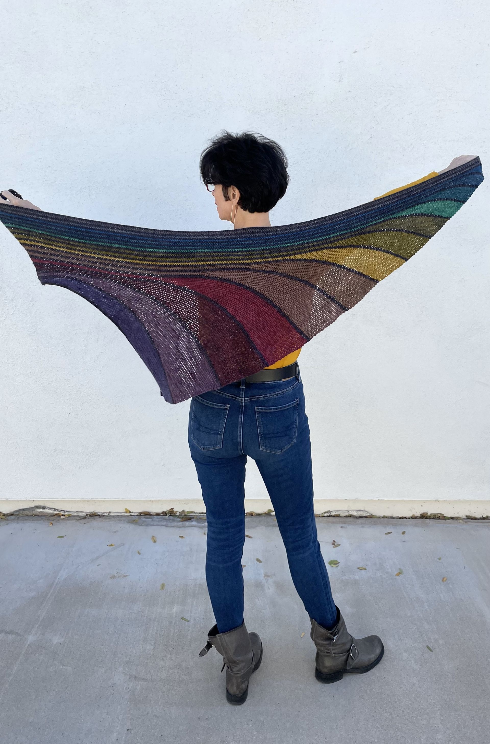 Woman in jeans holding up a rainbow-colored shawl against a white wall.