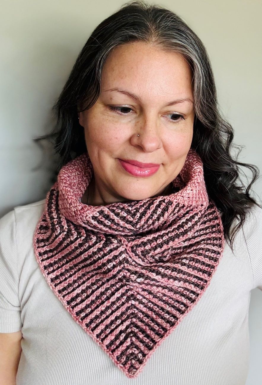 Woman wearing a pink and brown striped knit cowl. She's smiling, with gray hair, indoors.
