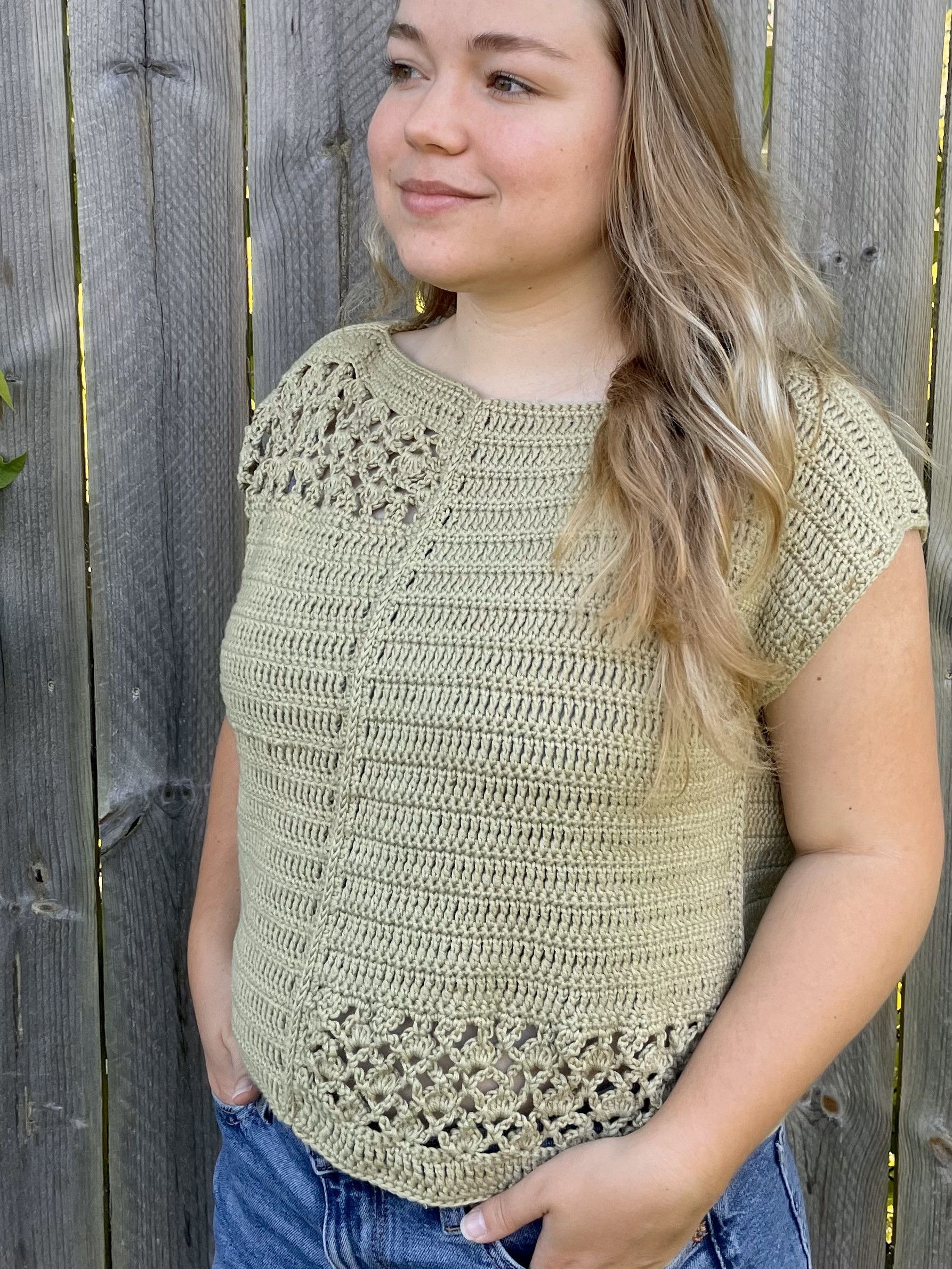 Woman in a light green crochet top smiles, standing outdoors. The top has a V-neck, short sleeves, and a textured pattern.