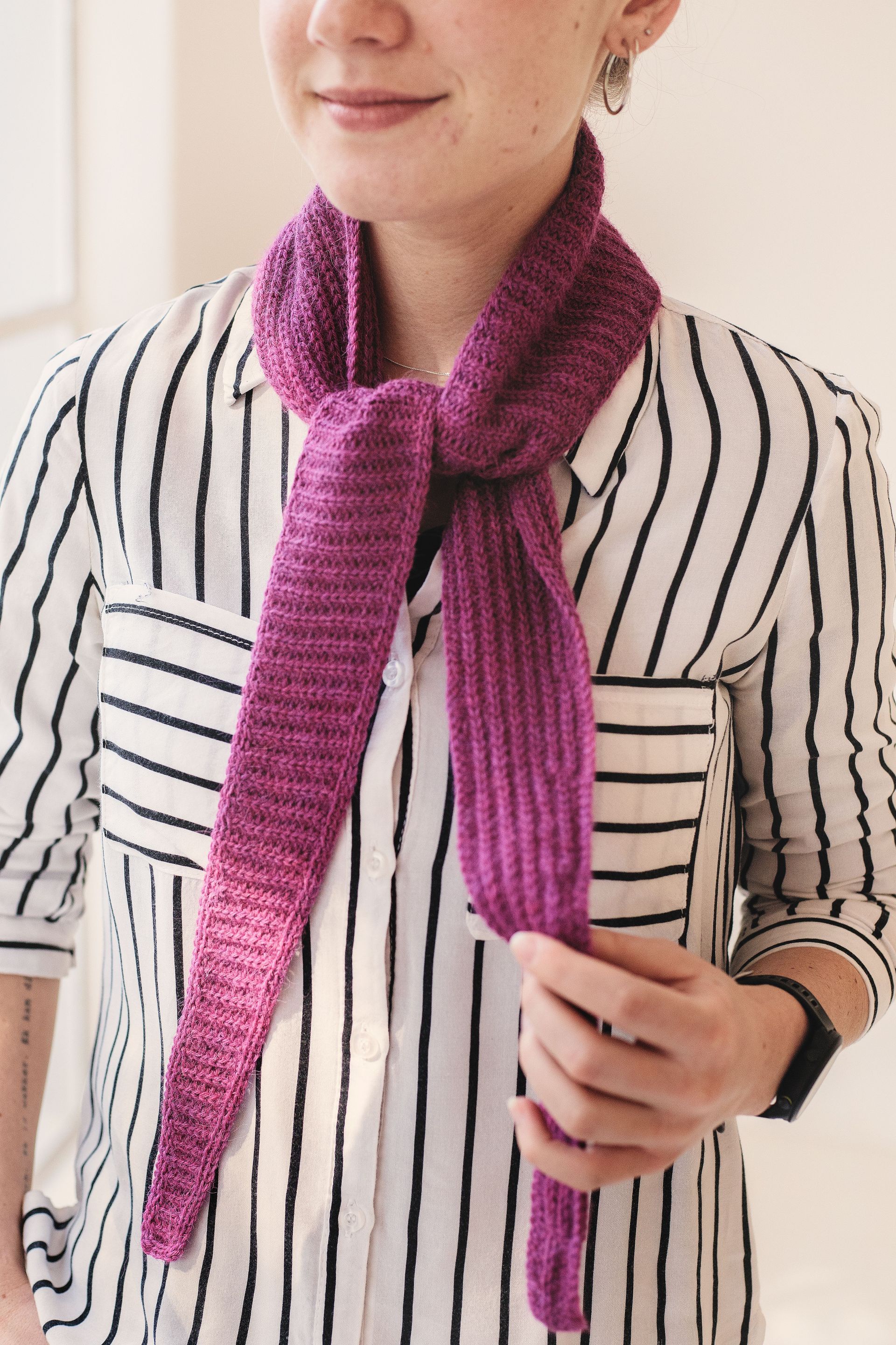 Woman wearing a purple crochetd scarf tied around her neck, standing in front of a window, wearing a striped shirt.