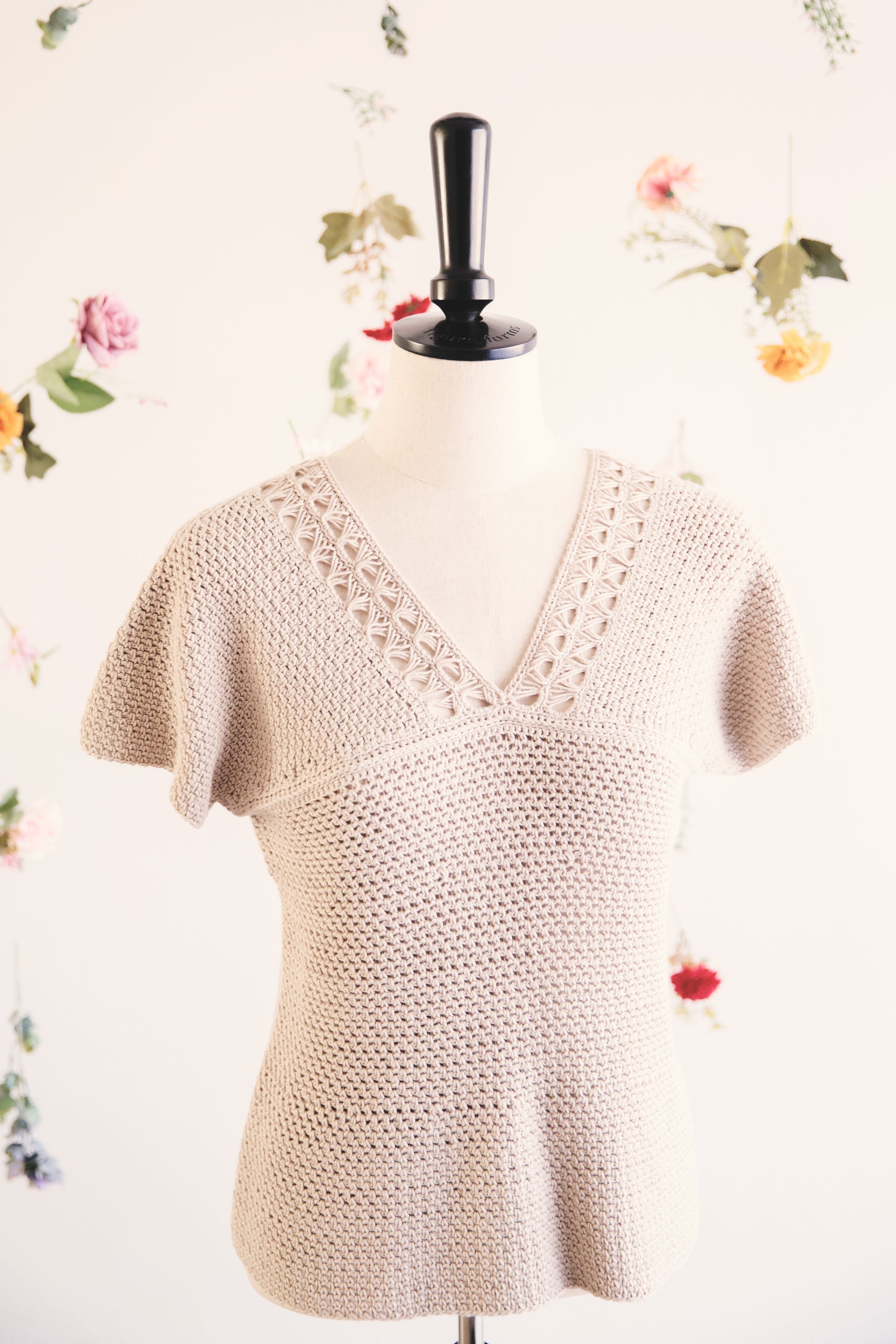 Beige crochet top on a mannequin with a V-neck, flared sleeves, and waist. Floral backdrop.