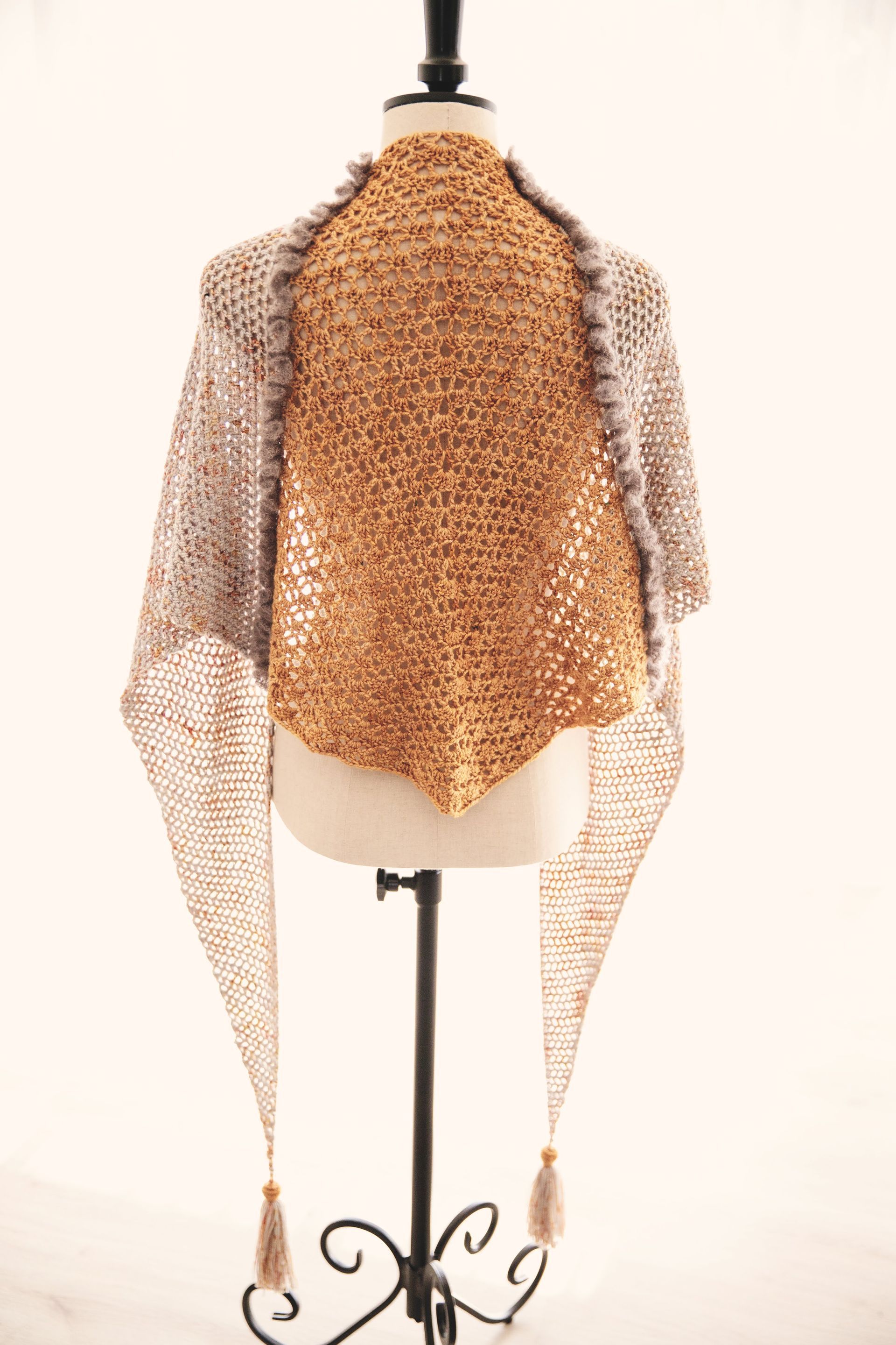 A crocheted shawl on a mannequin. Tan, brown, and gray with tassels.