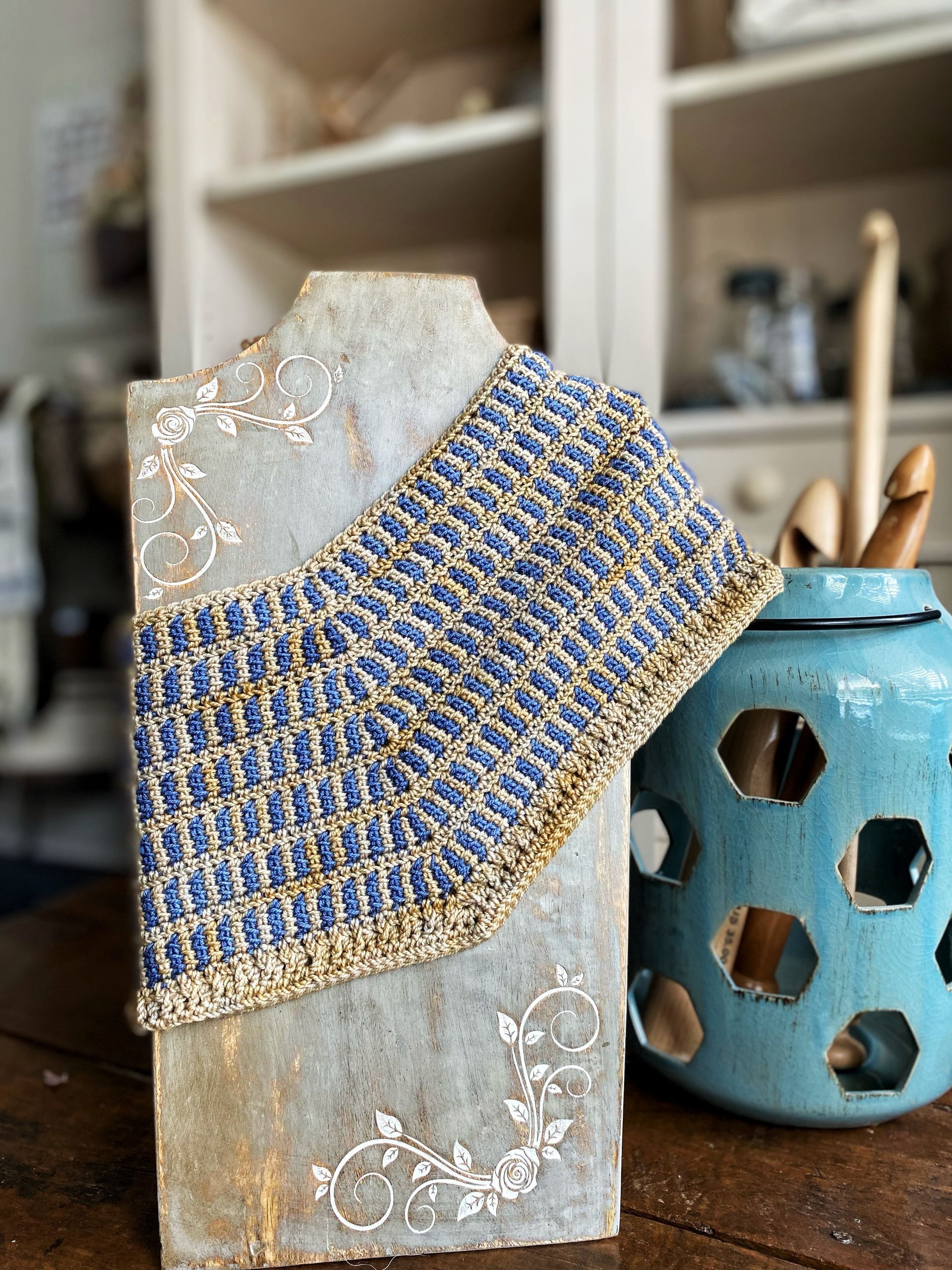 Crocheted blue and gold cowl displayed on a mannequin against a shelf.