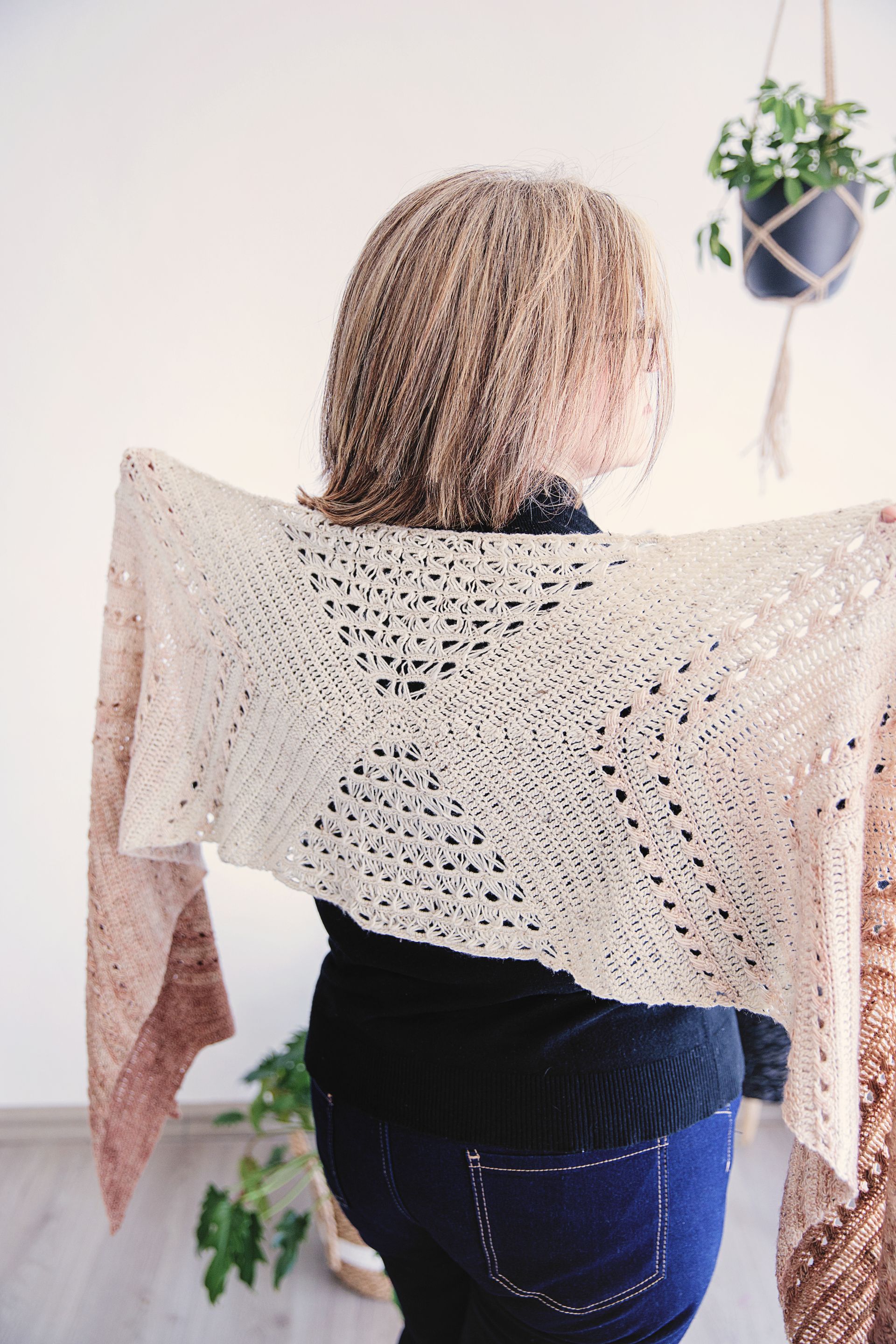 Woman wearing a cream and light brown crocheted shawl; indoors, plants in background.
