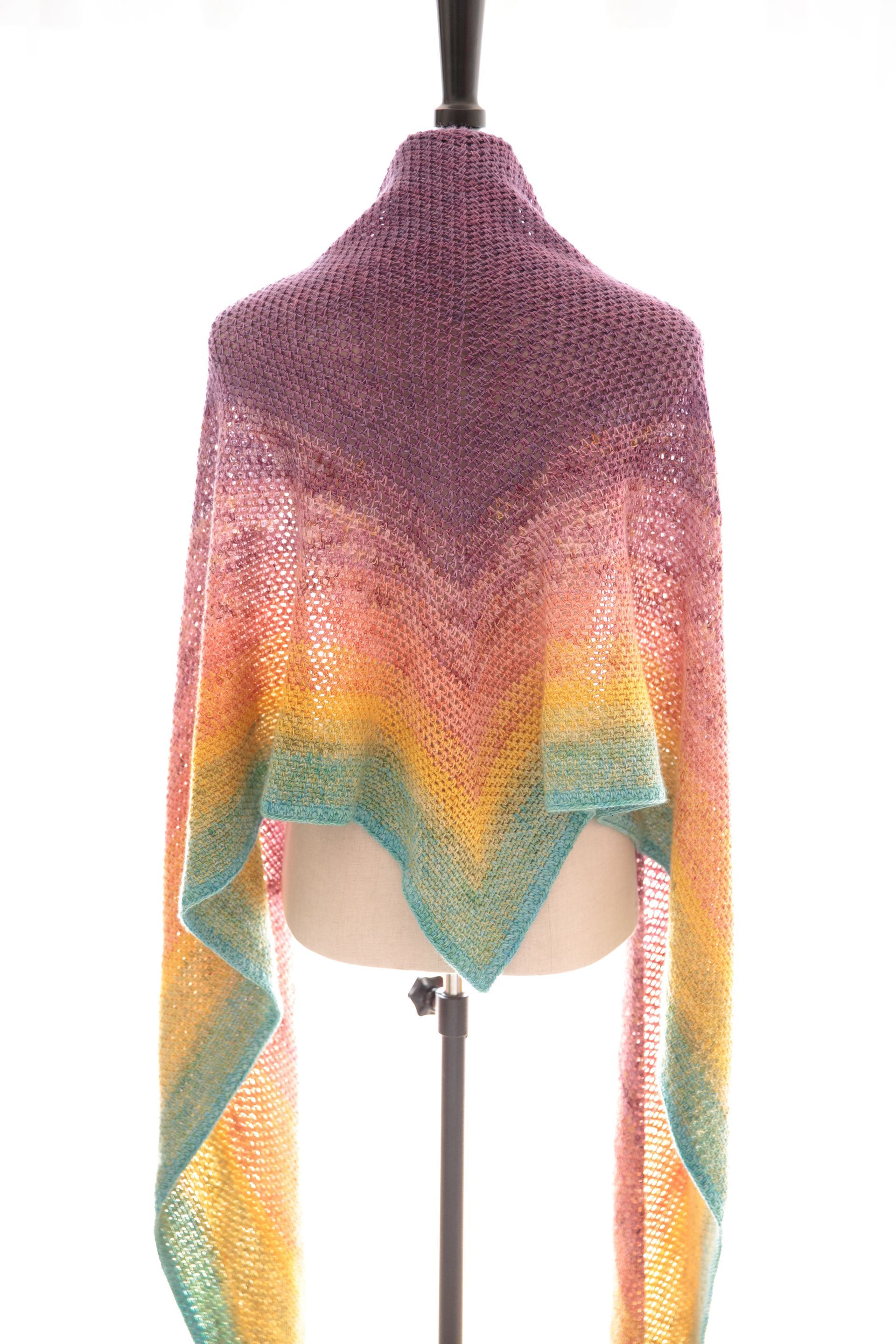 A colorful, triangular shawl in a gradient of pink, orange, yellow, and teal, draped on a mannequin.