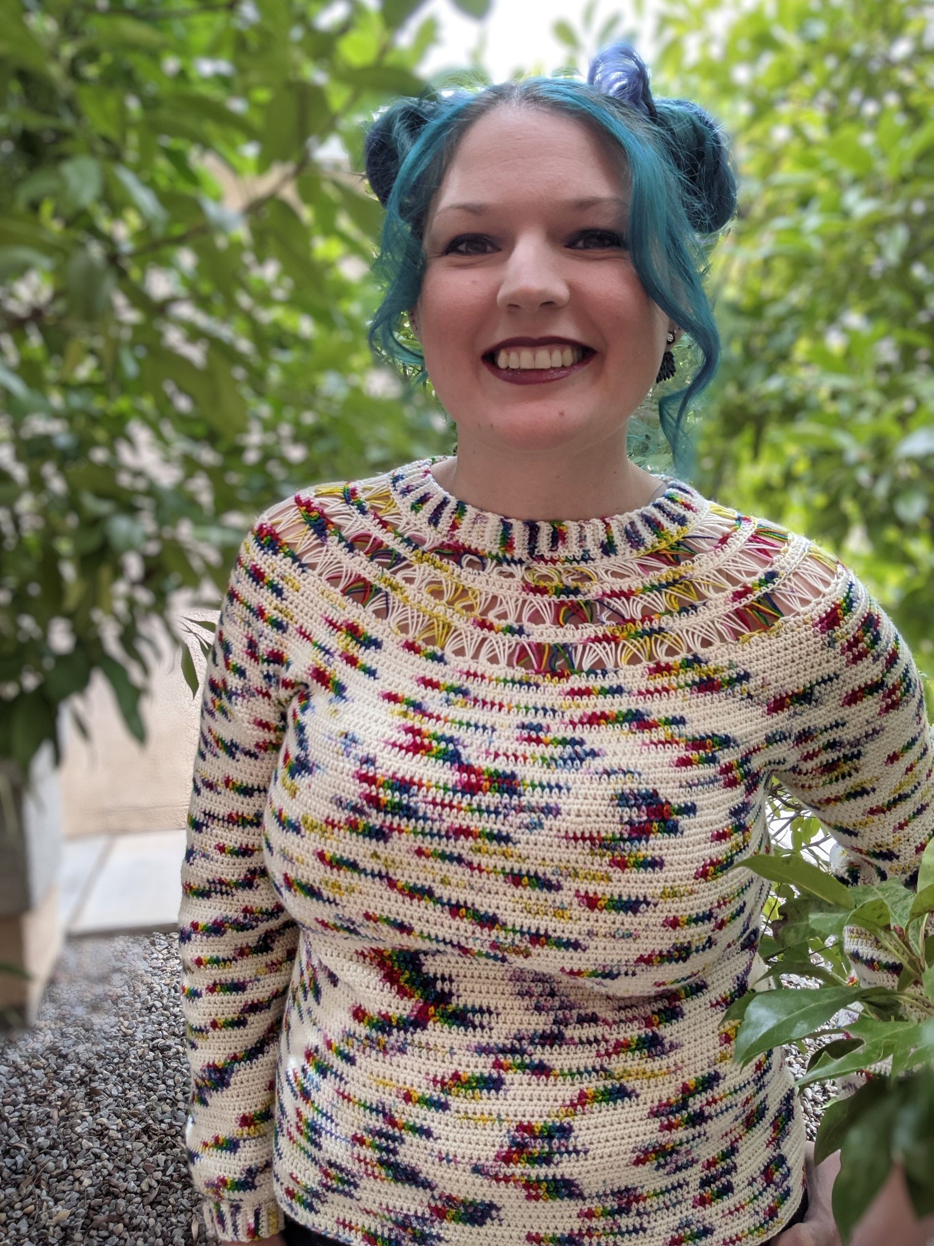 Woman with teal space buns smiling, wearing a colorful speckled sweater outdoors.
