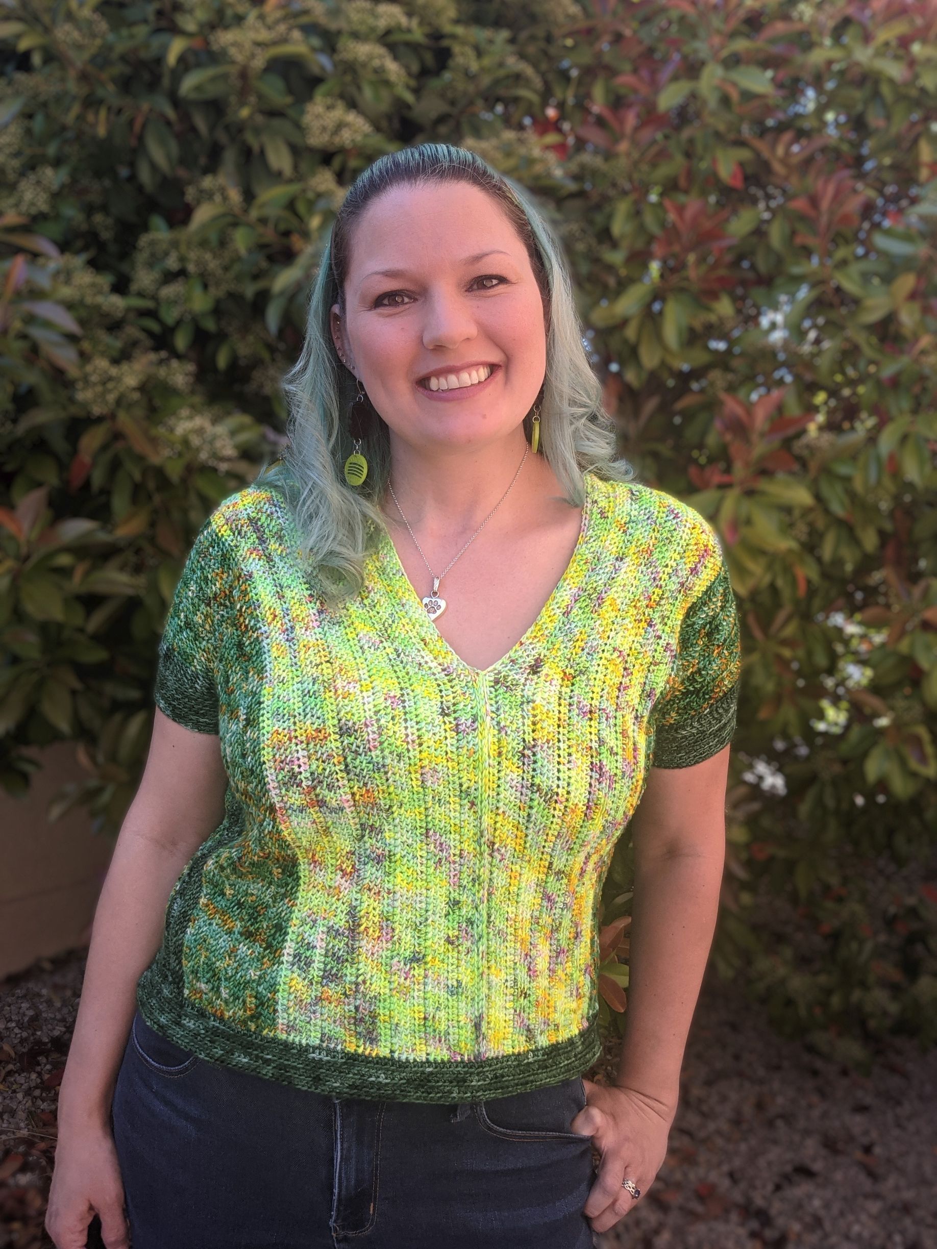 Woman with turquoise hair smiles, wearing a green and yellow knitted shirt, standing outdoors.
