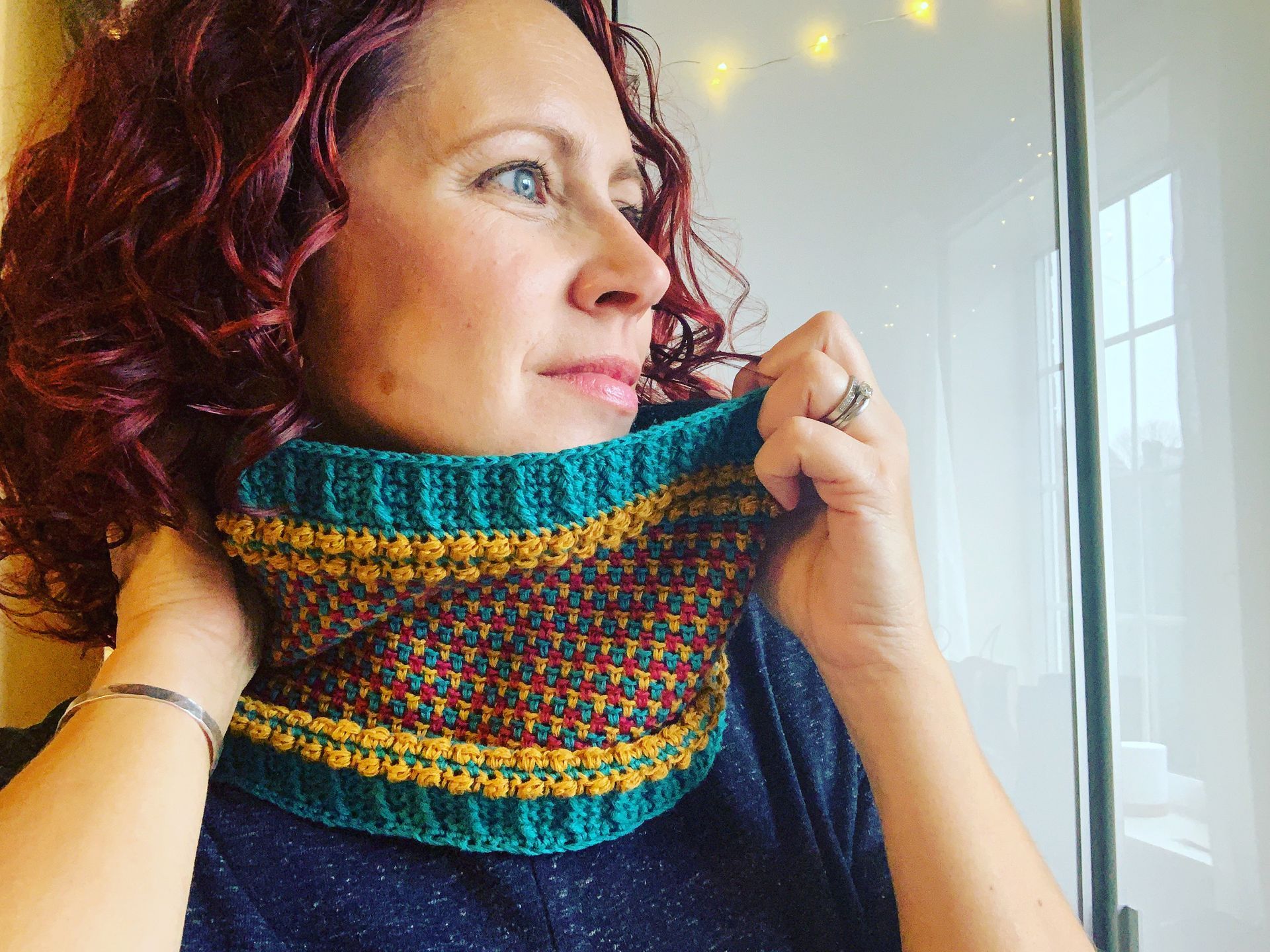 Woman with curly red hair wearing a turquoise and gold crocheted cowl. She is smiling.