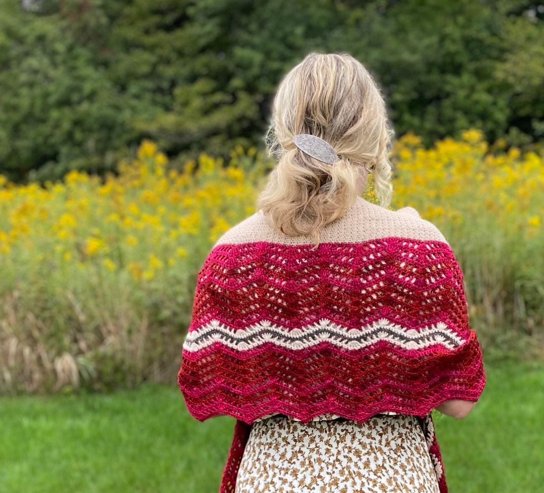 Woman in red and cream shawl outdoors, hair pulled back with clip, standing in front of yellow flowers.