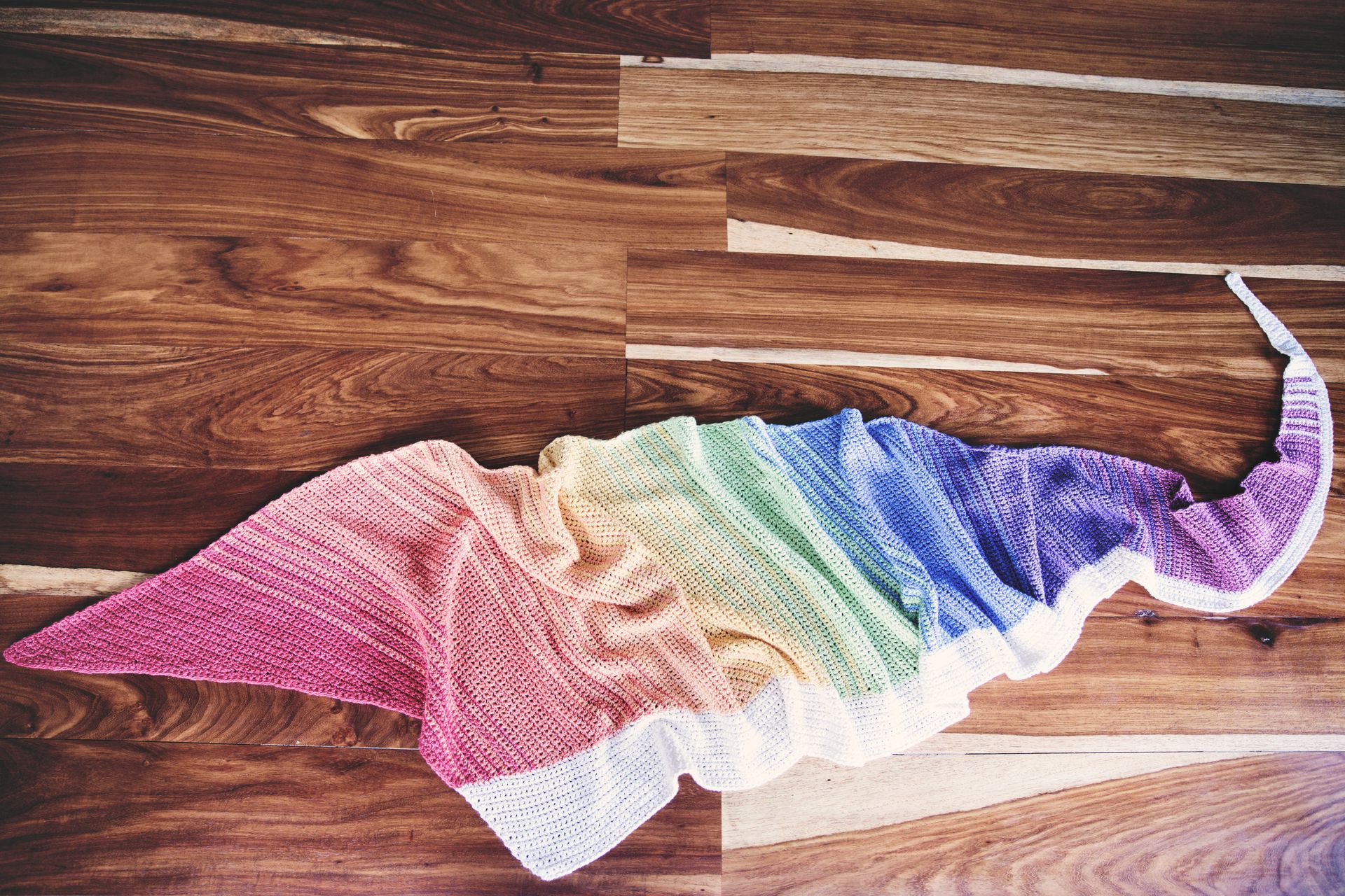 Crocheted rainbow shawl on a wooden floor, colors blend from pink to purple and white.