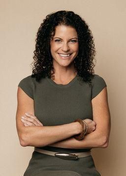 A smiling woman with dark curly hair, wearing a dark green top and a belt, posing with arms crossed against a tan background.