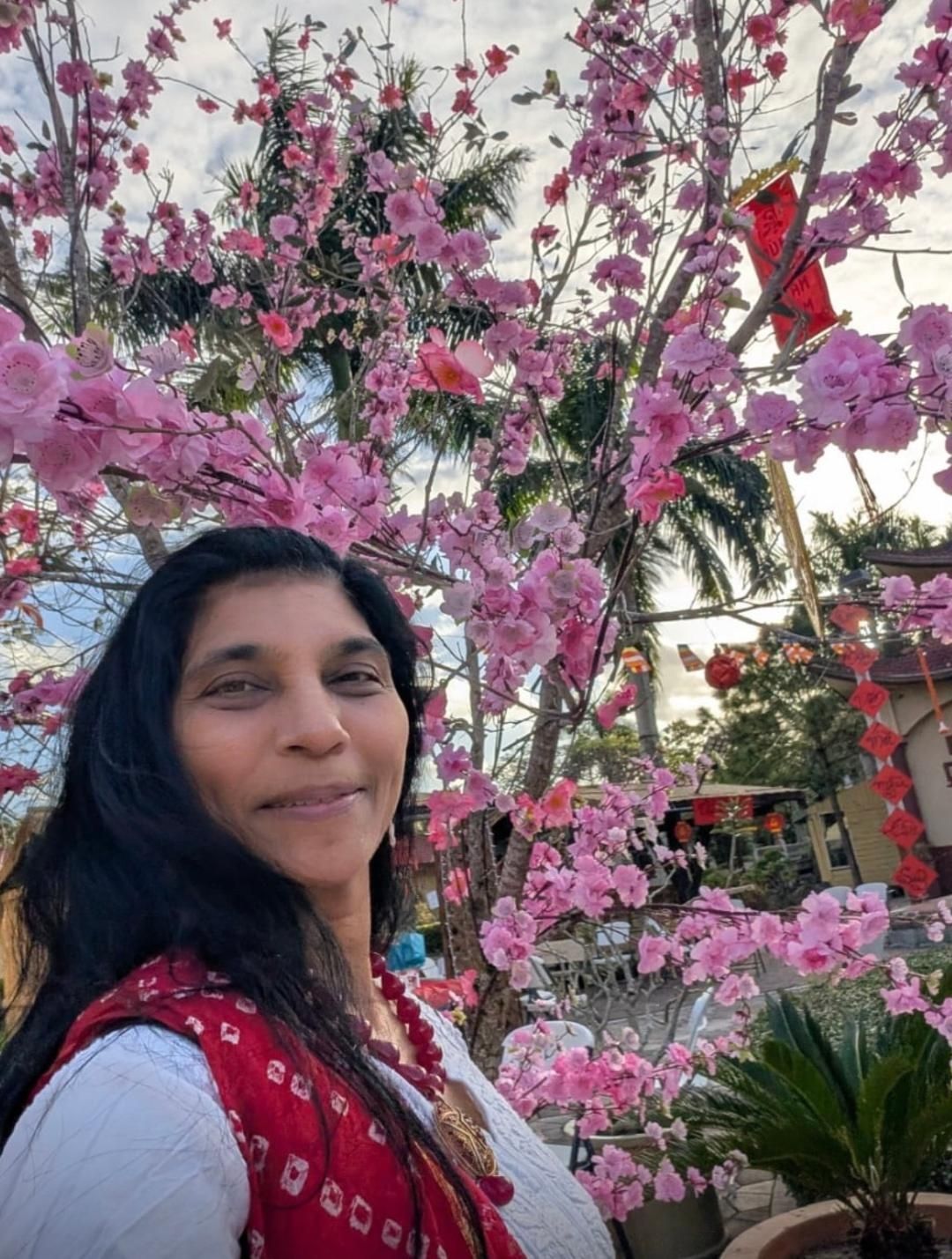 A person smiling in front of a tree decorated with vibrant pink blossoms during a sunny outdoor setting.