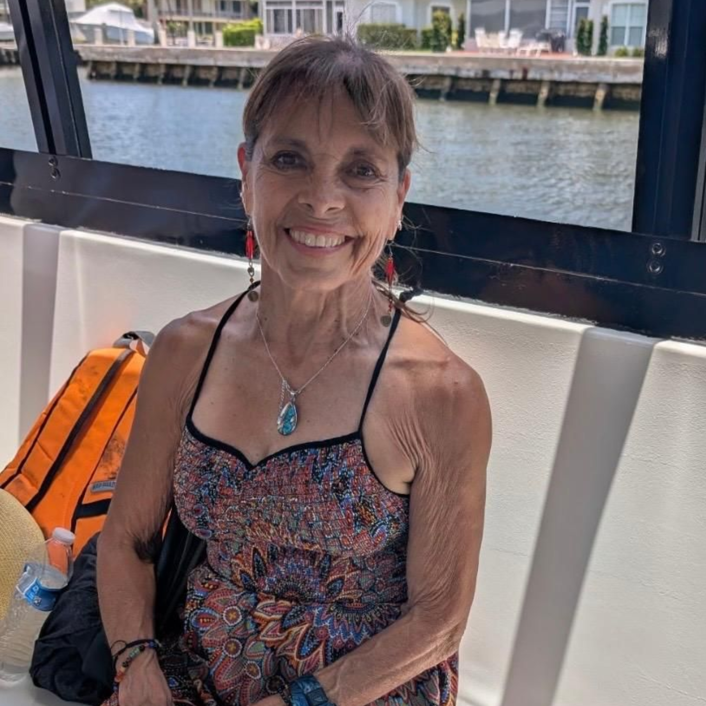 A smiling person sits on a boat bench, wearing a patterned summer dress, with waterfront houses in the background.