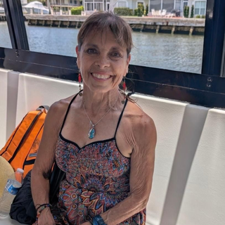 A smiling person sits on a boat bench, wearing a patterned summer dress, with waterfront houses in the background.