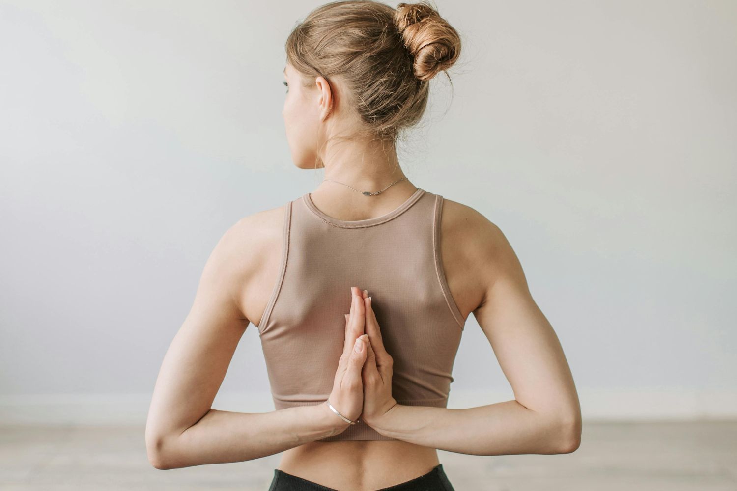 Woman in a beige tank top, with hands clasped behind back, performing yoga pose. Against a white wall.