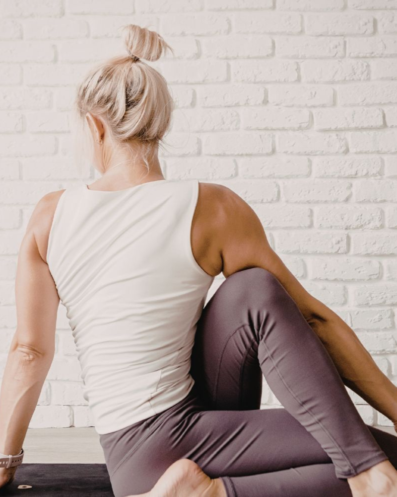 Woman in yoga pose, white top, purple leggings, twisting torso, seated, against white brick wall.