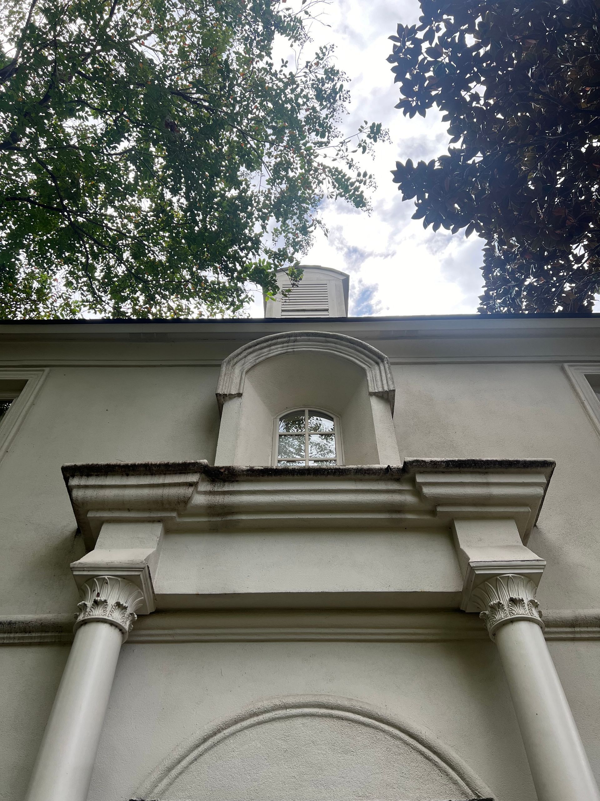 White building facade with classical columns, arched window, and tree canopy against a cloudy sky.