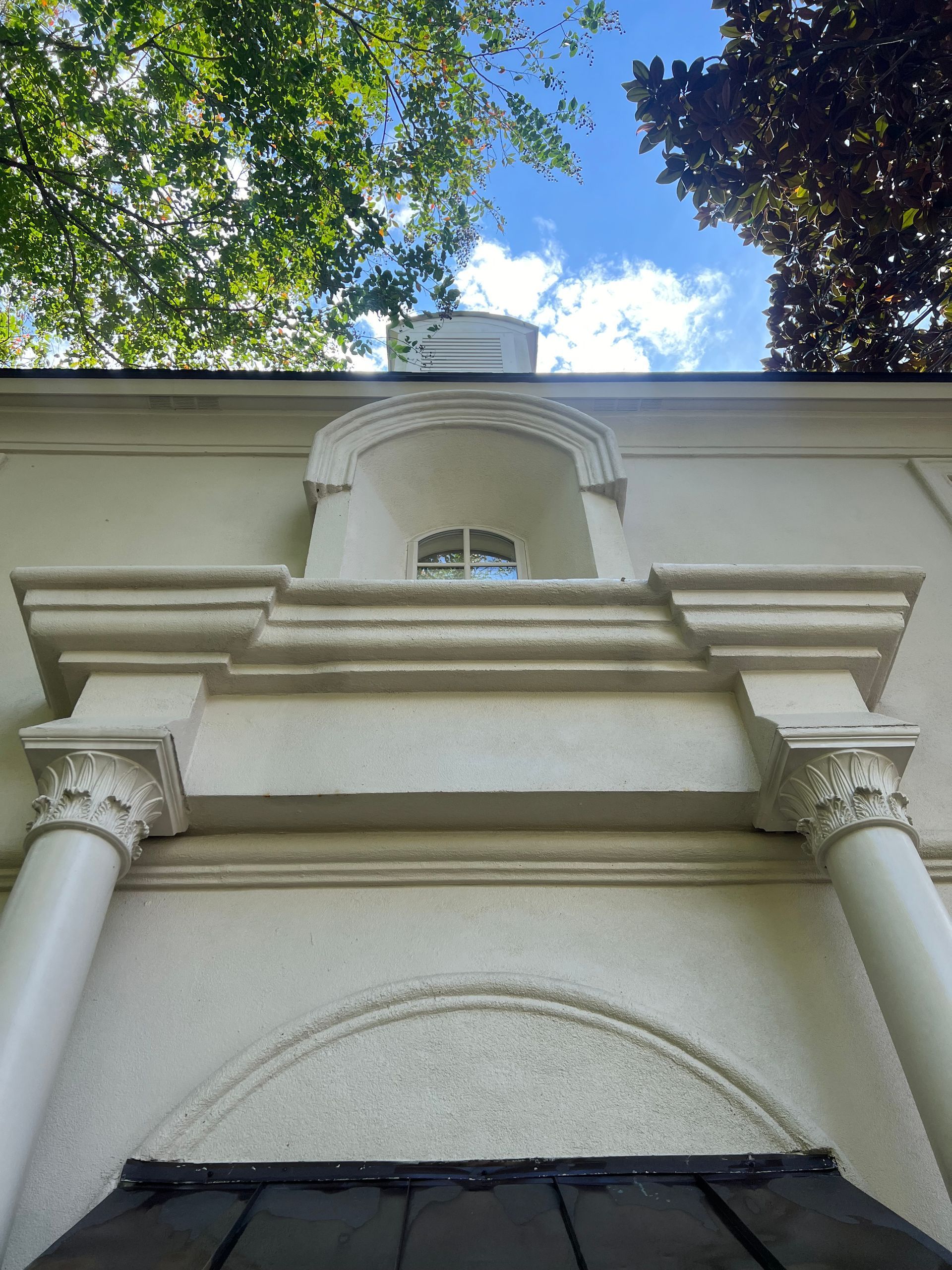 White stucco building with a decorative facade, arched window, and columns against a blue sky with trees.