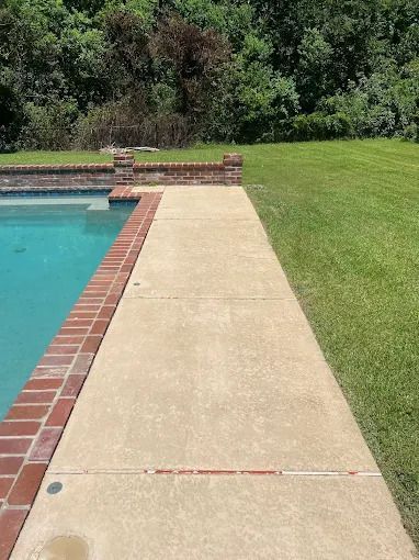 Concrete walkway alongside a swimming pool with brick edging, next to a grassy lawn.