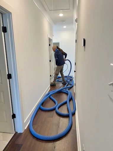 Person vacuuming hallway floor with blue hose. Wooden floor, white walls, black doors.