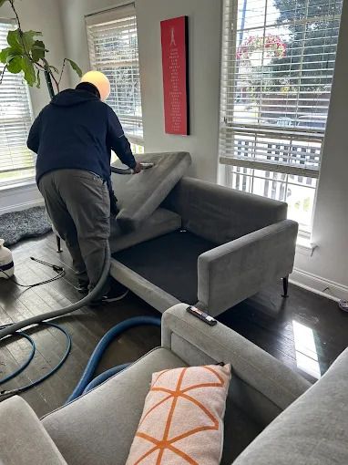Person cleaning a gray couch with a vacuum hose in a well-lit room with large windows.