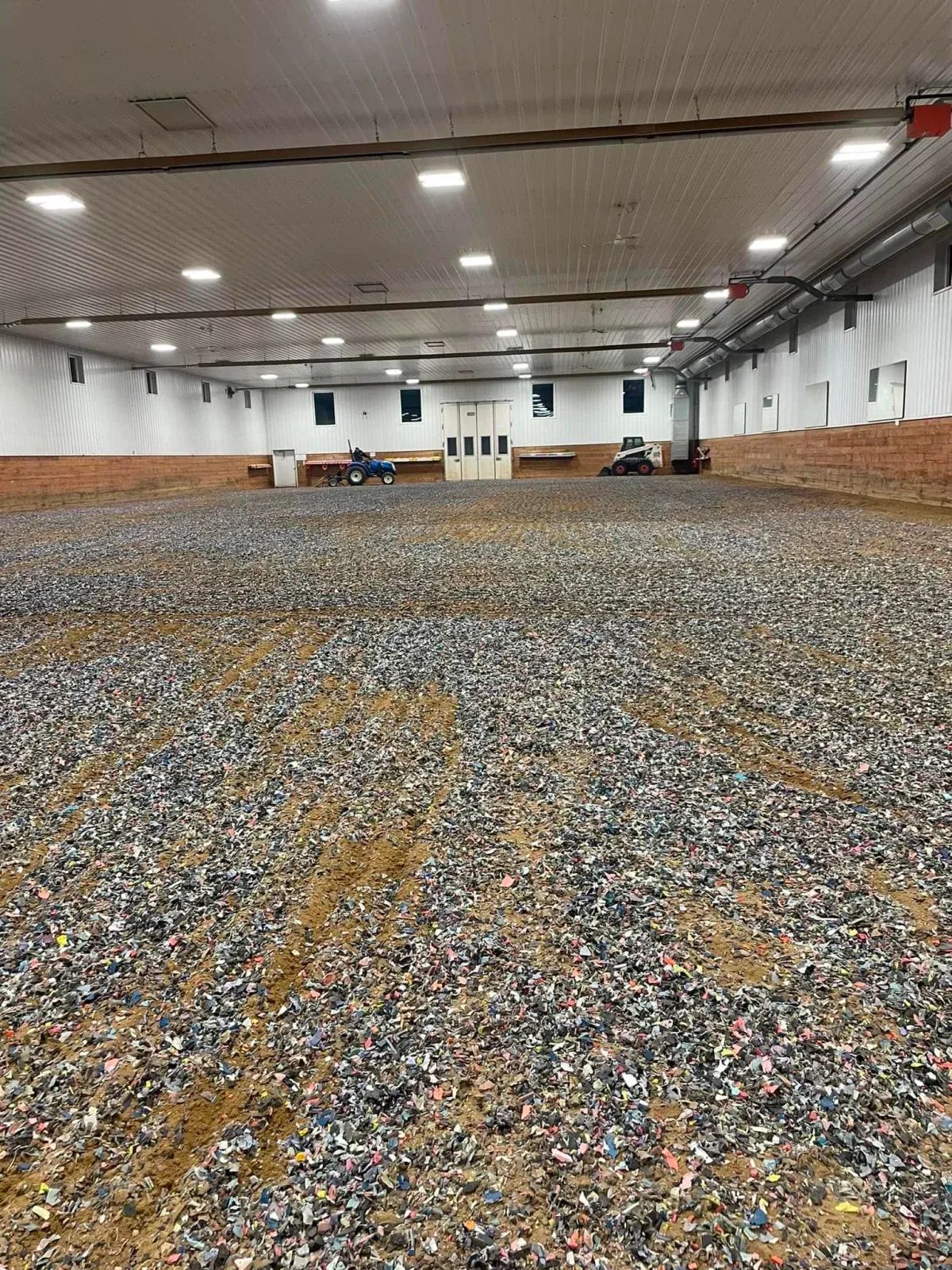 An indoor riding arena filled with footing. White walls, overhead lighting, and a safety net are visible.