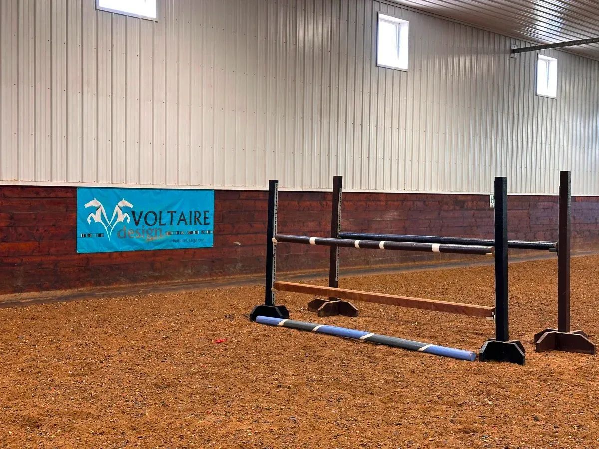 Indoor equestrian arena with a jump set up. A blue Voltaire sign is on the wall, and the ground is covered in wood chips.