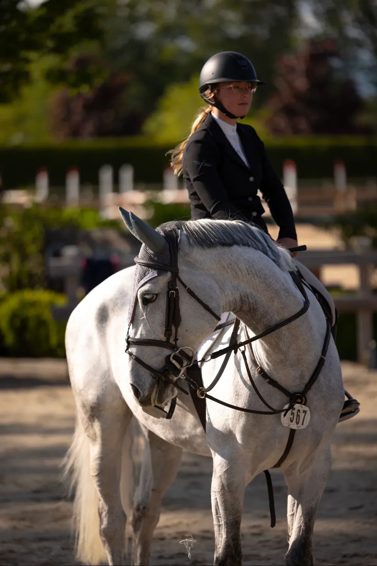 Young woman on a gray horse at a competition, wearing a black helmet and jacket. They are in an outdoor arena.