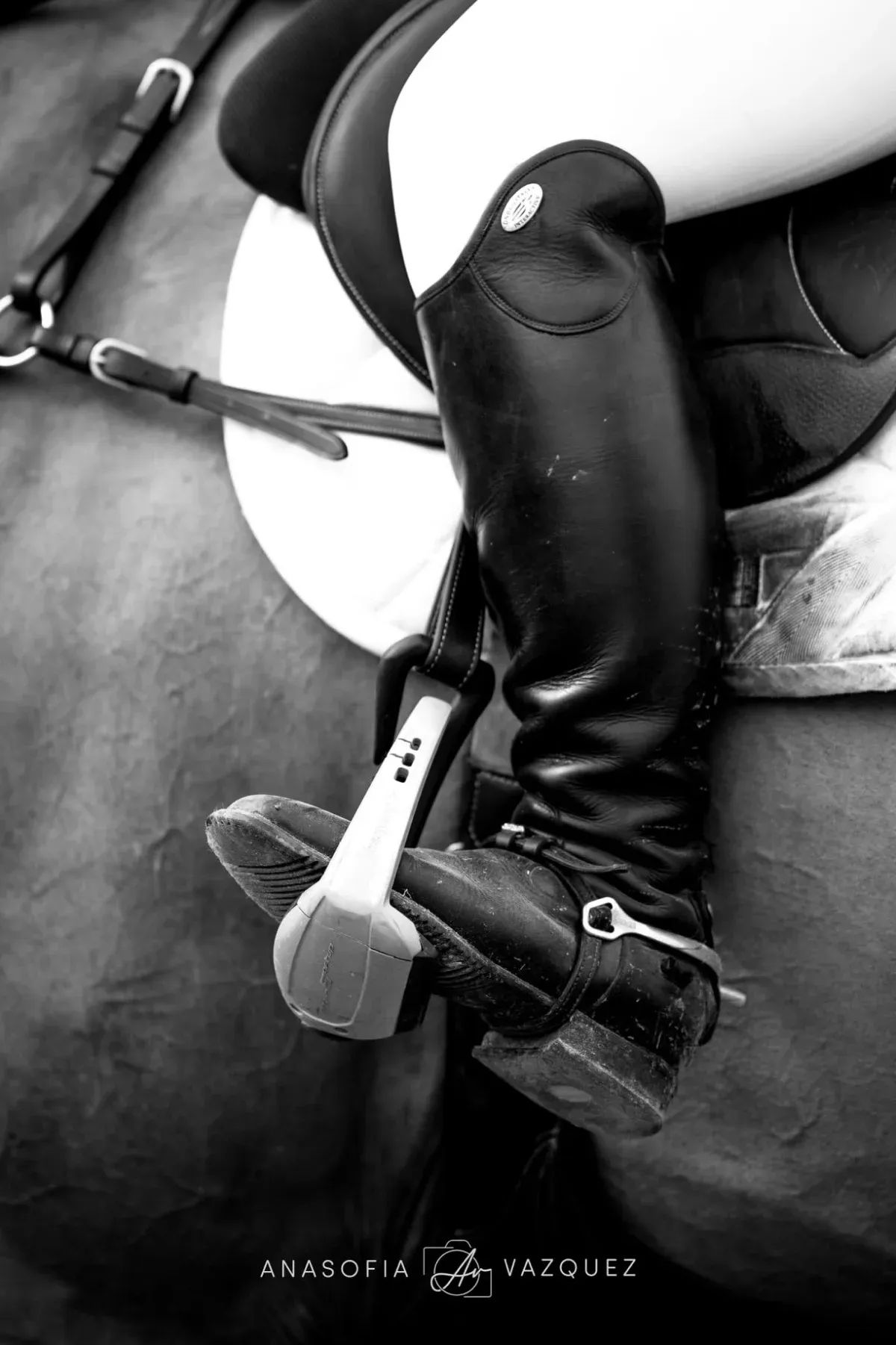Black and white photo of a rider's leg in a tall black riding boot with a spur, foot in the stirrup, sitting on a horse.