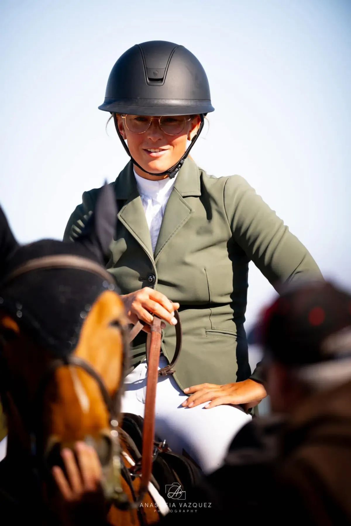 A woman in riding attire sits on a horse, smiling. She wears a dark helmet and green jacket.