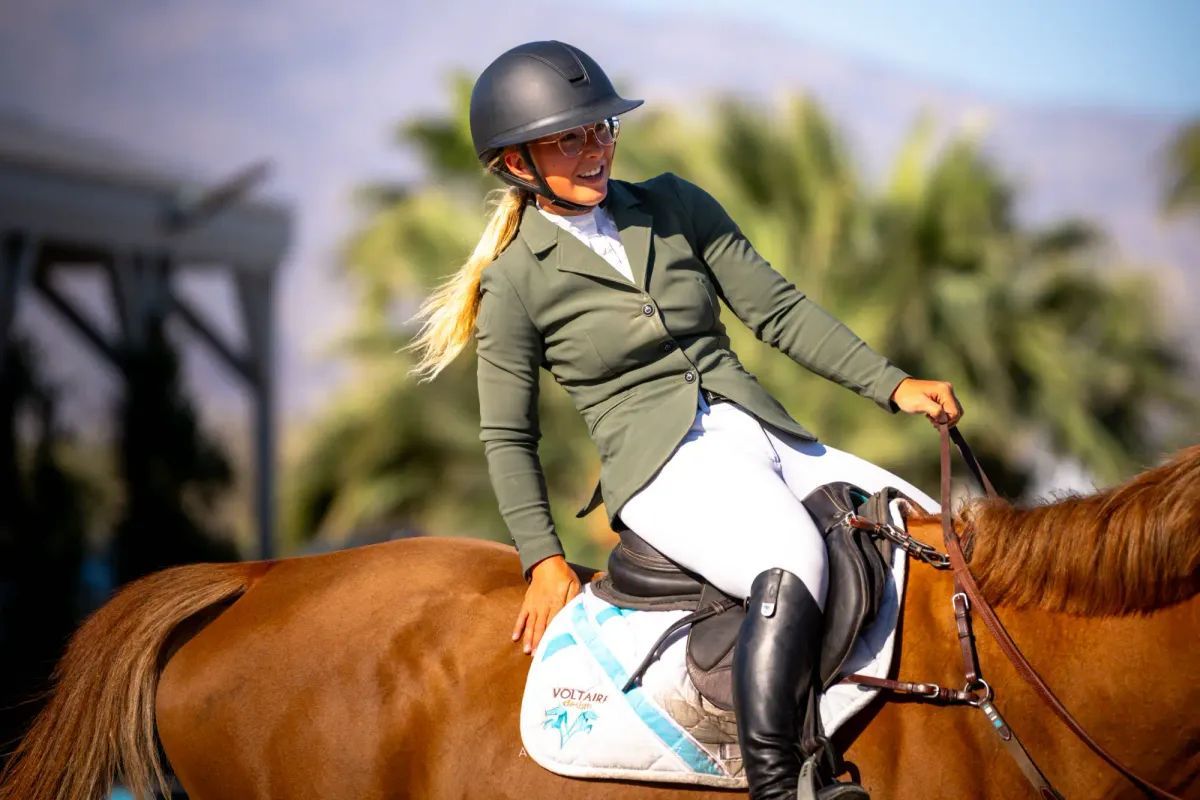 A blonde equestrian in a green jacket and black helmet smiles while riding a brown horse.