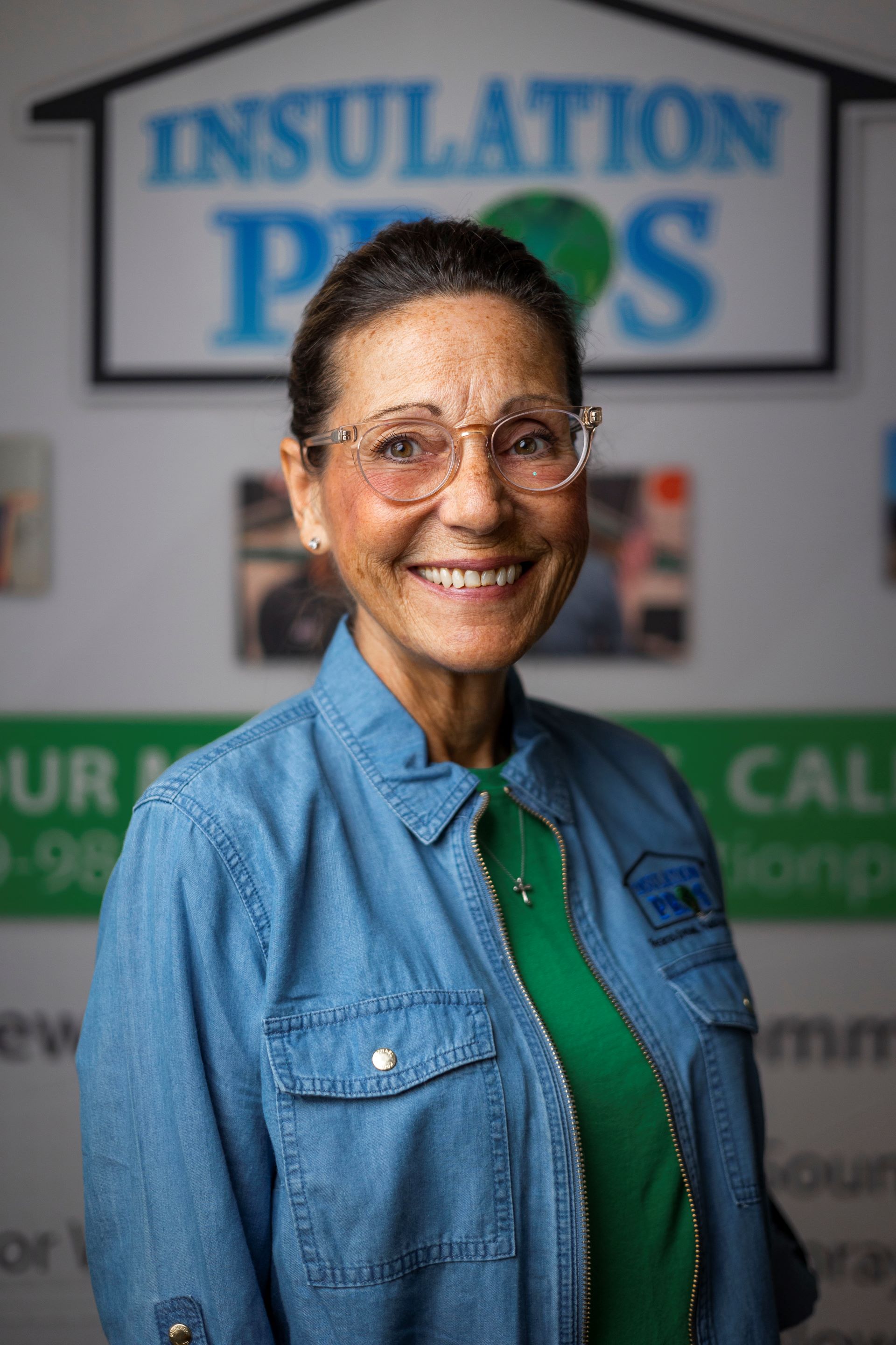 A woman wearing glasses and a denim shirt is smiling in front of a sign for insulation pros.