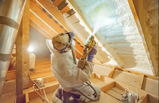 A man is spraying insulation on the ceiling of an attic.